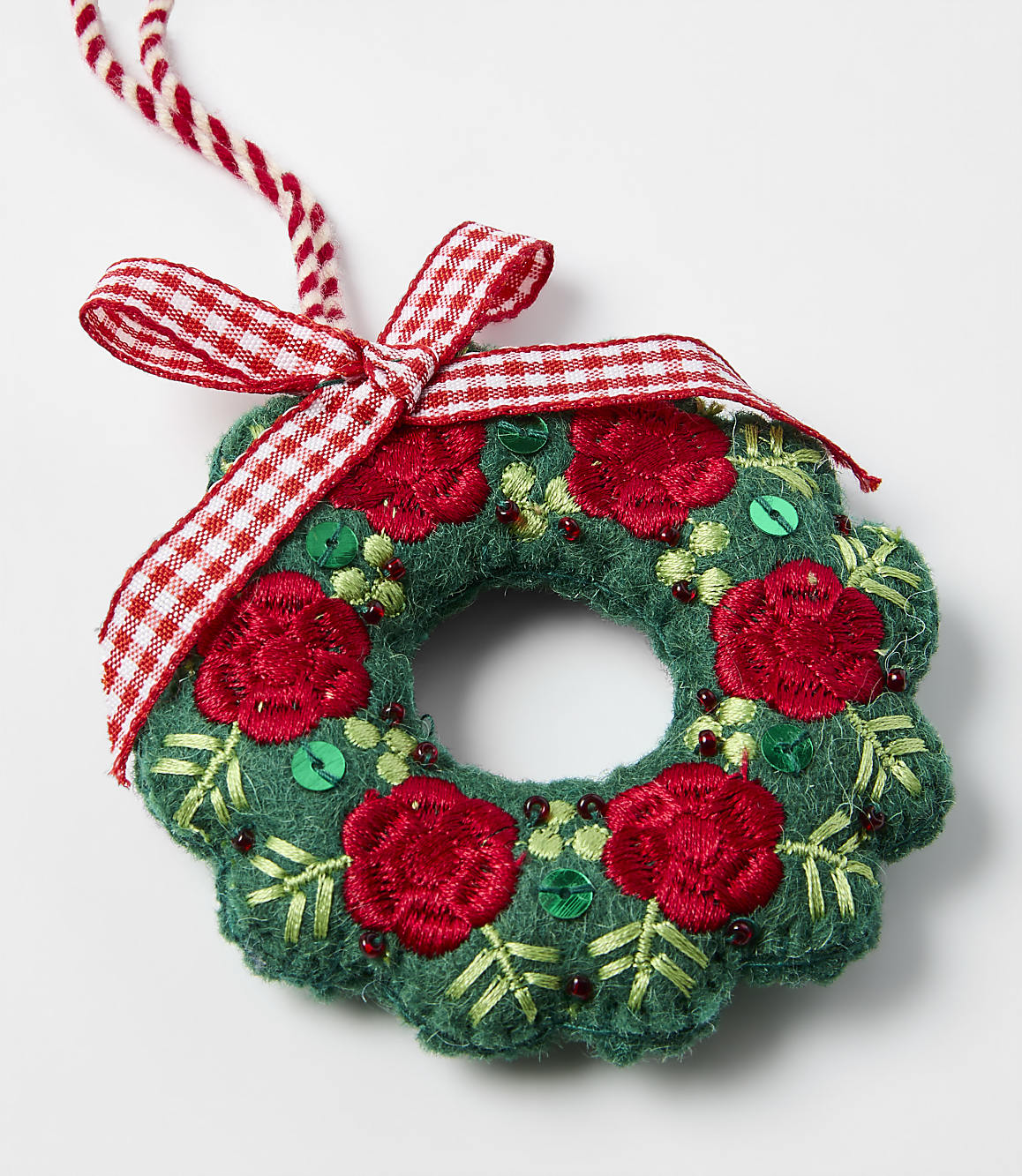 A green fabric wreath adorned with red embroidered flowers and a red-and-white checkered bow rests on a plain white background.