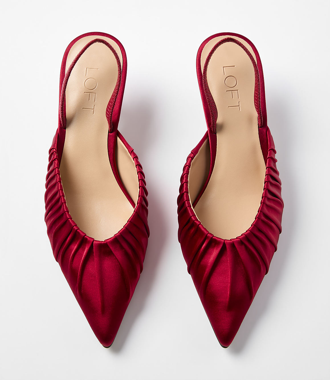 A pair of red satin pointed-toe mules with ruched detailing sits against a plain white background.