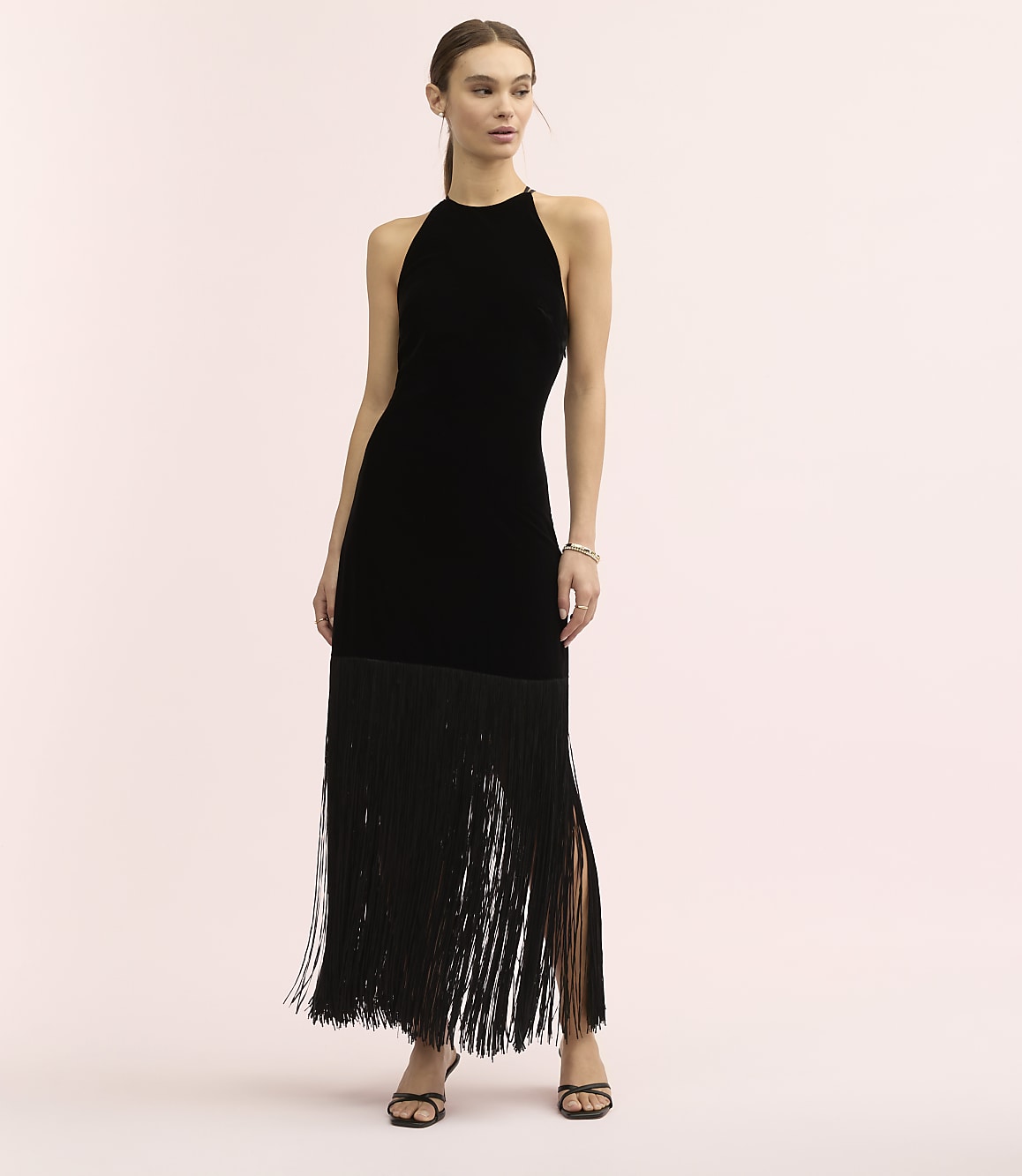 A woman wearing a black velvet halter dress with a long fringe skirt stands against a plain light pink background.