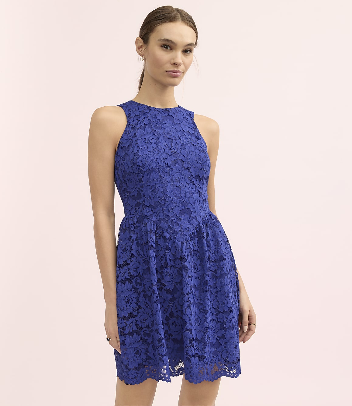 A woman wearing a royal blue lace dress stands against a plain light pink background.