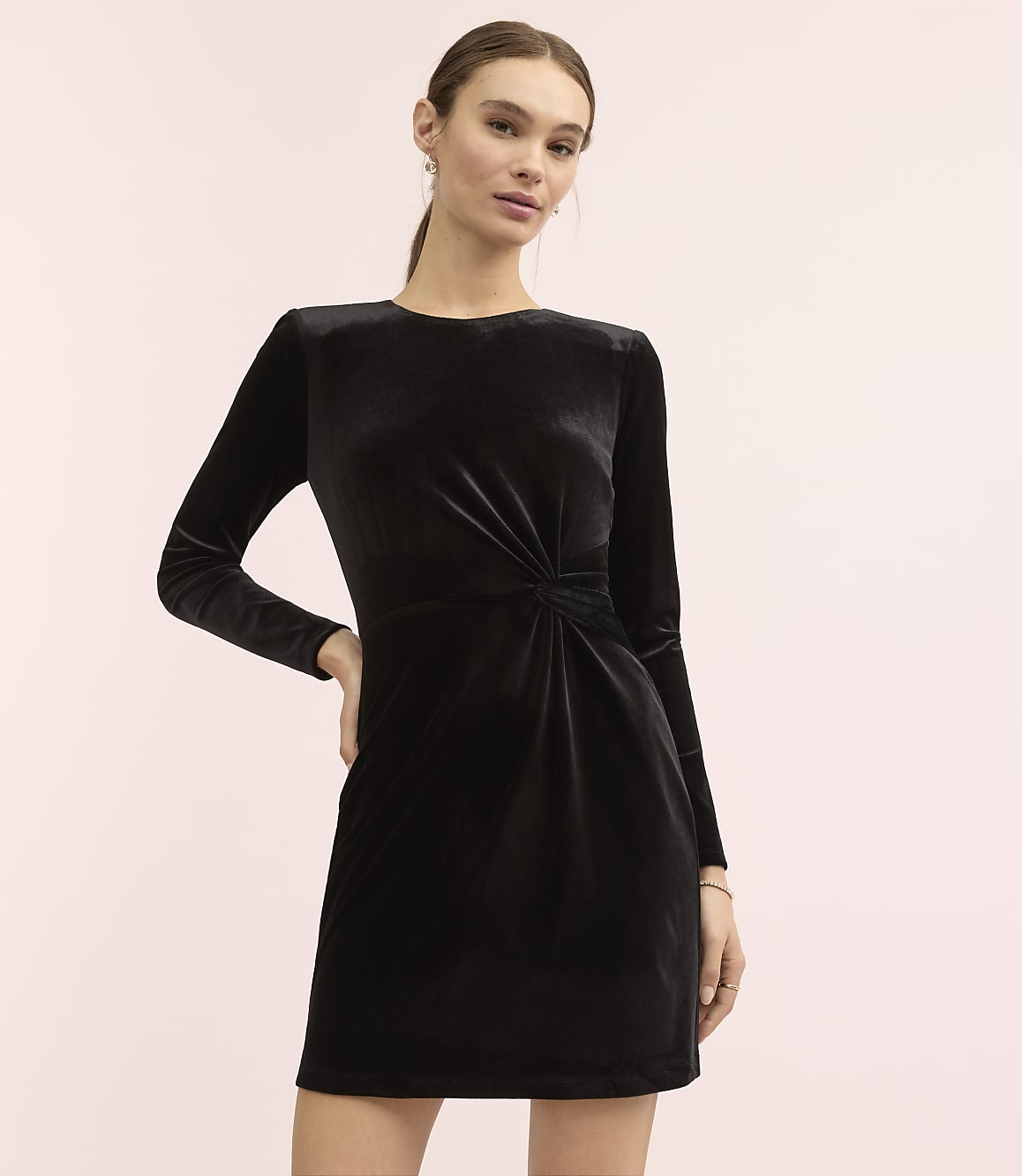 A woman wearing a long-sleeved black velvet dress stands against a plain light pink background.