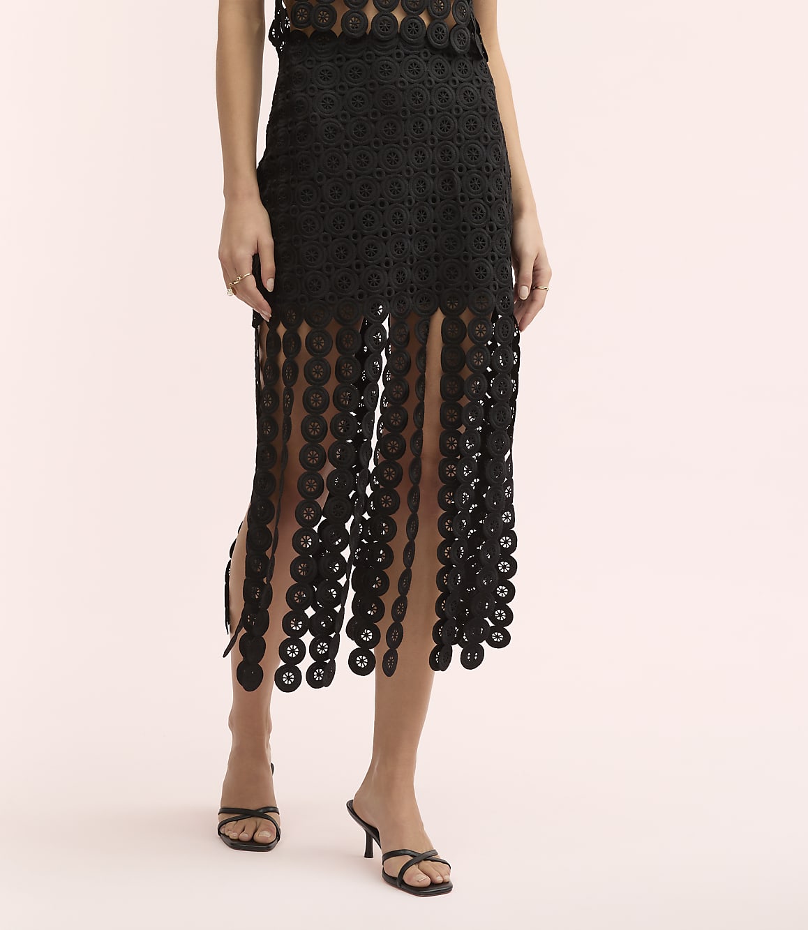 A person wears a black lace-patterned dress and black sandals against a plain light pink background.