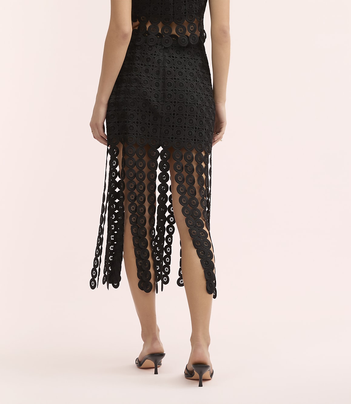 A woman stands with her back to the camera wearing a black lace skirt and heels against a plain light pink background.