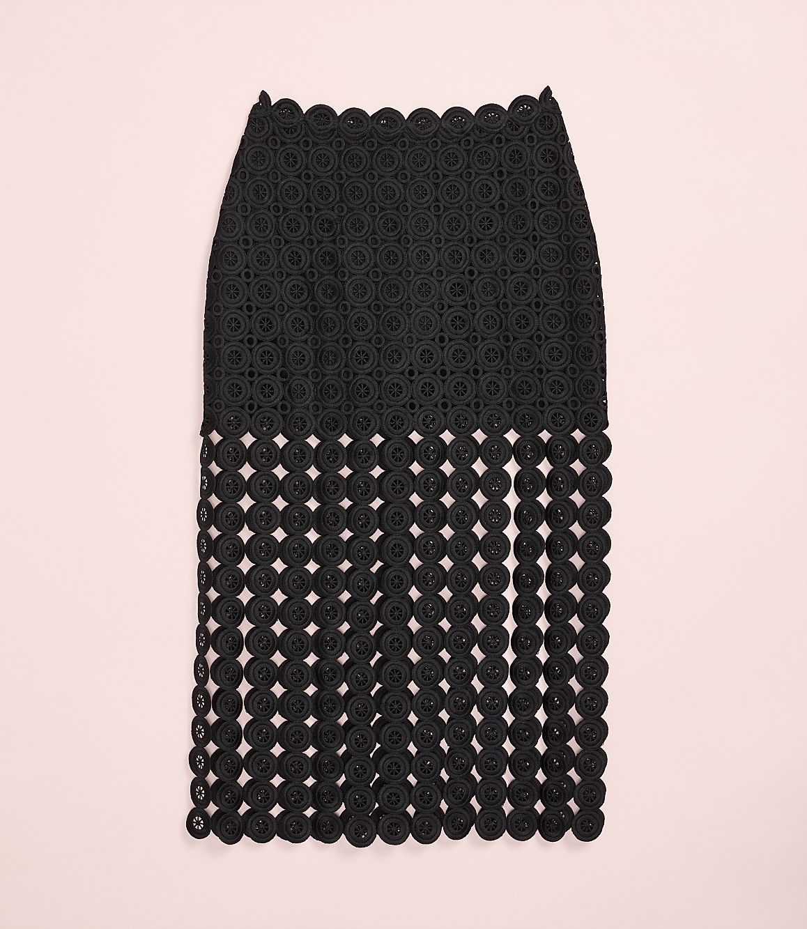 A black, intricately patterned skirt with a fringed bottom hangs against a plain light pink background.