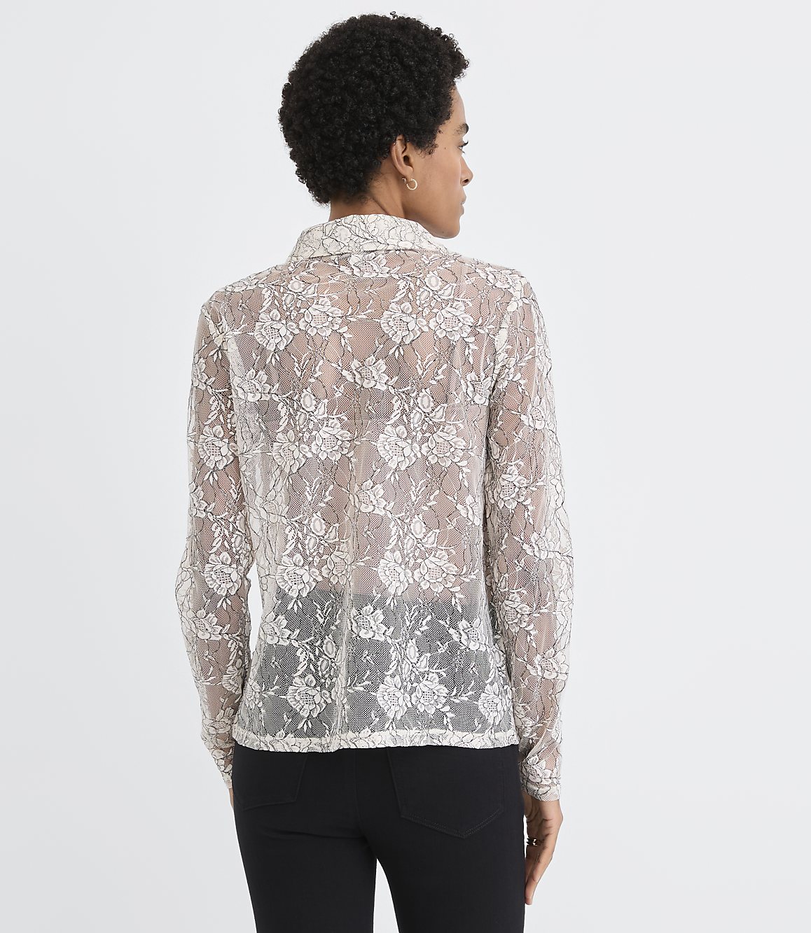 A woman wearing a sheer, floral lace blouse and black pants stands with her back to the camera against a plain white background.
