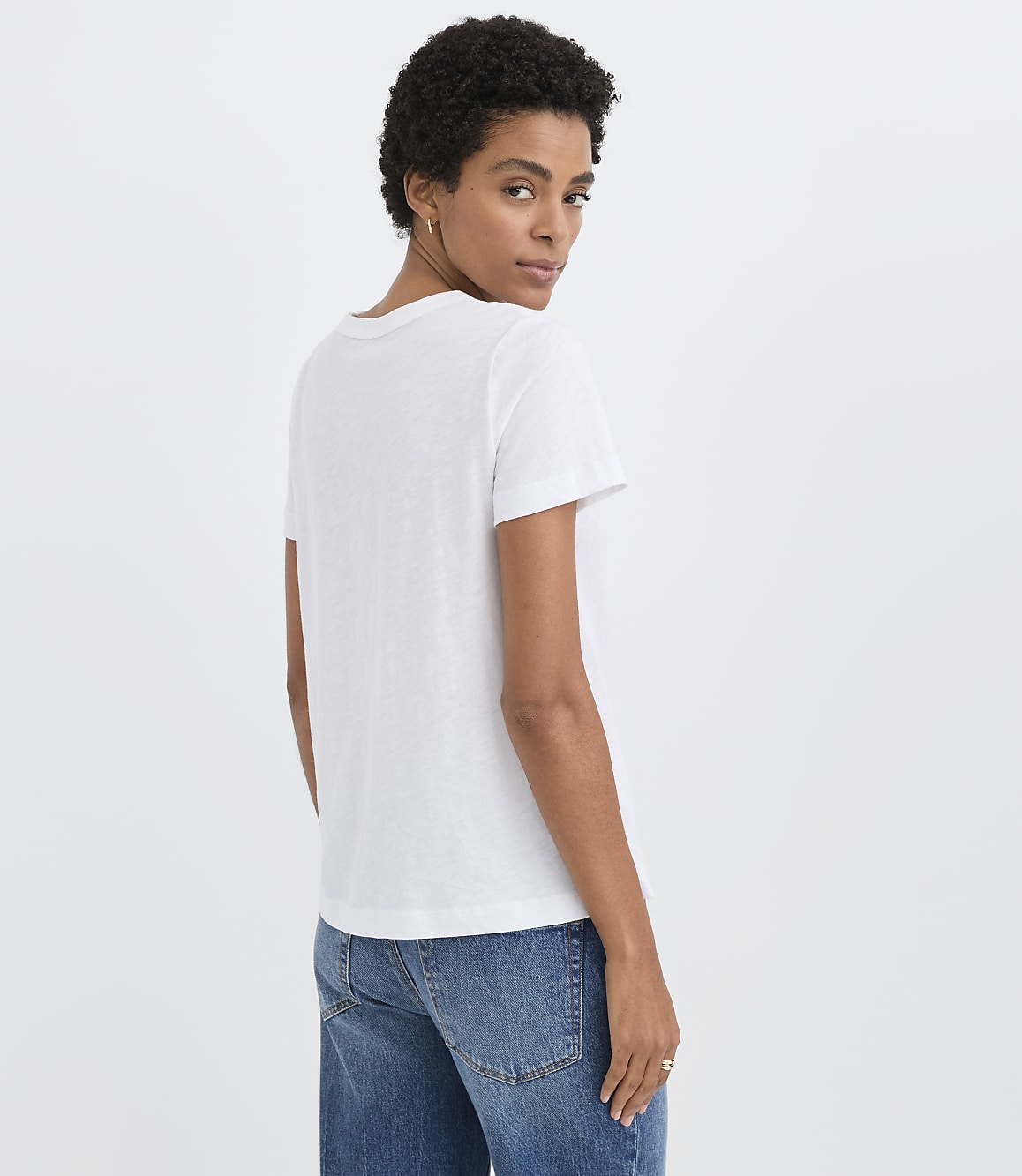 A woman wearing a white t-shirt and blue jeans looks over her shoulder against a plain white background.