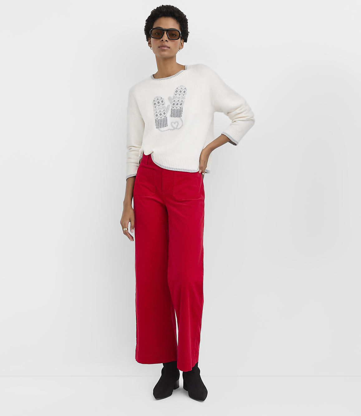 A woman wearing a white mitten-patterned sweater and red wide-leg pants stands against a plain white background.