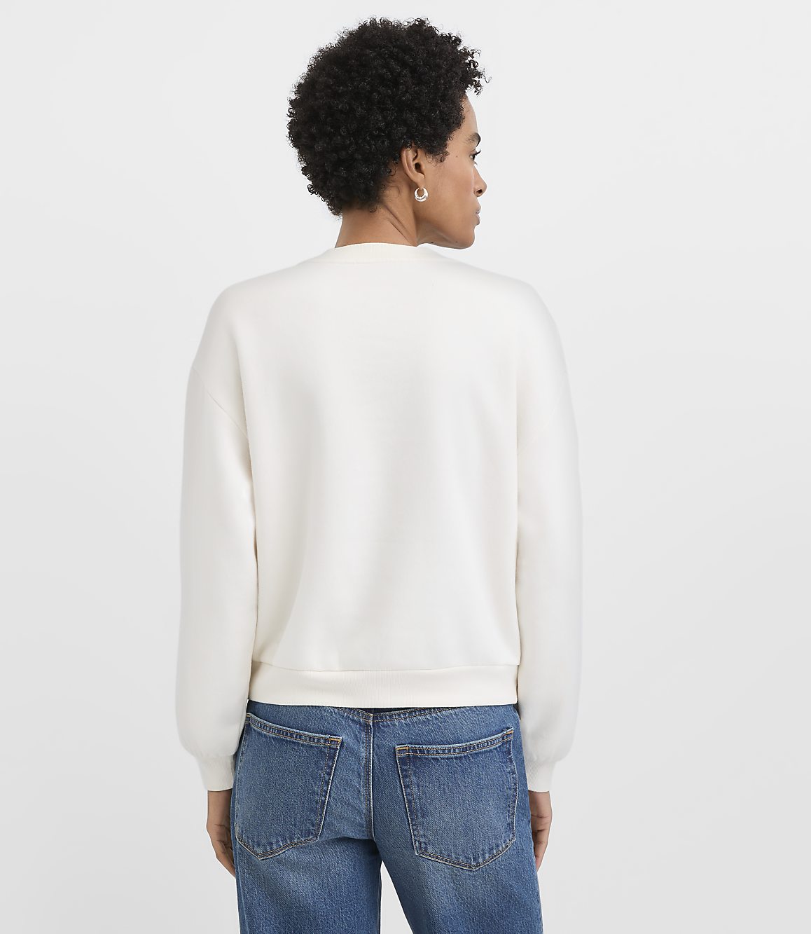A woman with curly hair wearing a white sweatshirt and blue jeans stands with her back to the camera against a plain white background.