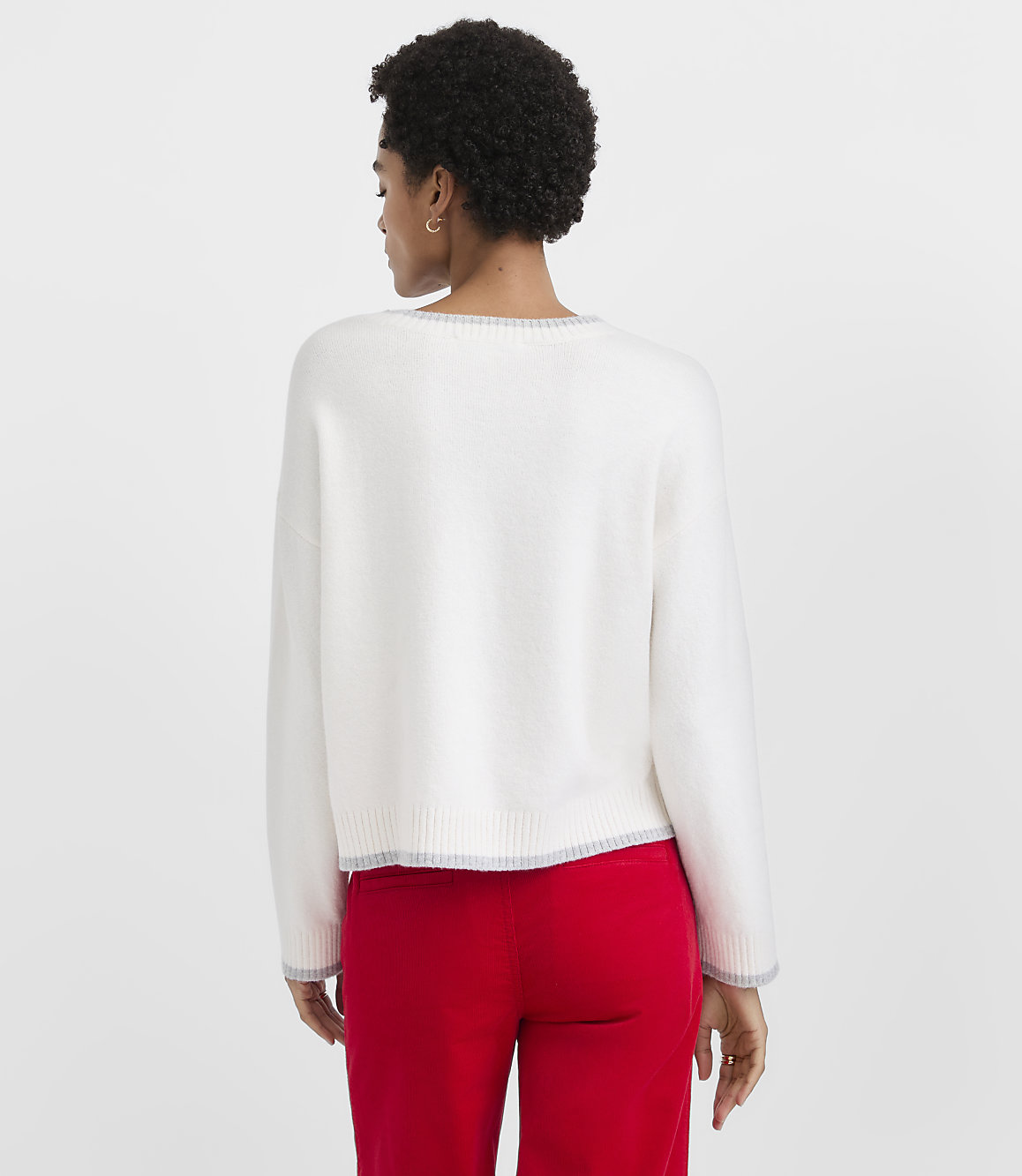 A woman with short curly hair wearing a white sweater and red pants stands against a plain light gray background.