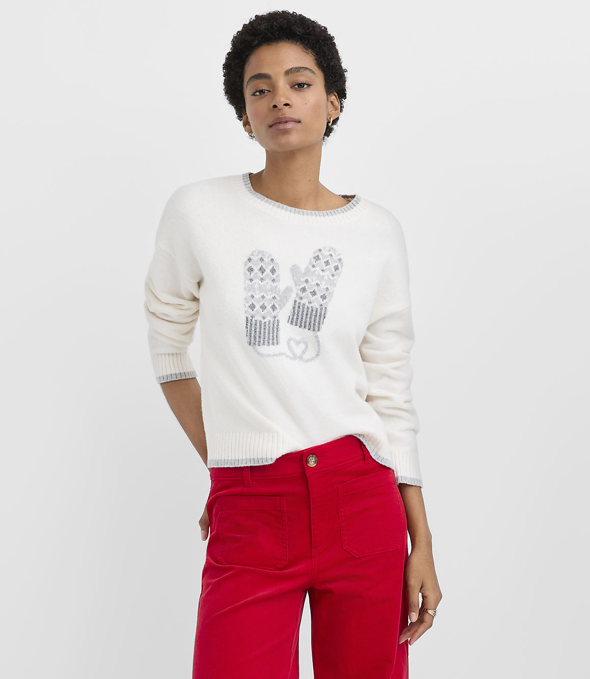 A woman wearing a white sweater with a mitten design and red pants stands against a plain white background.