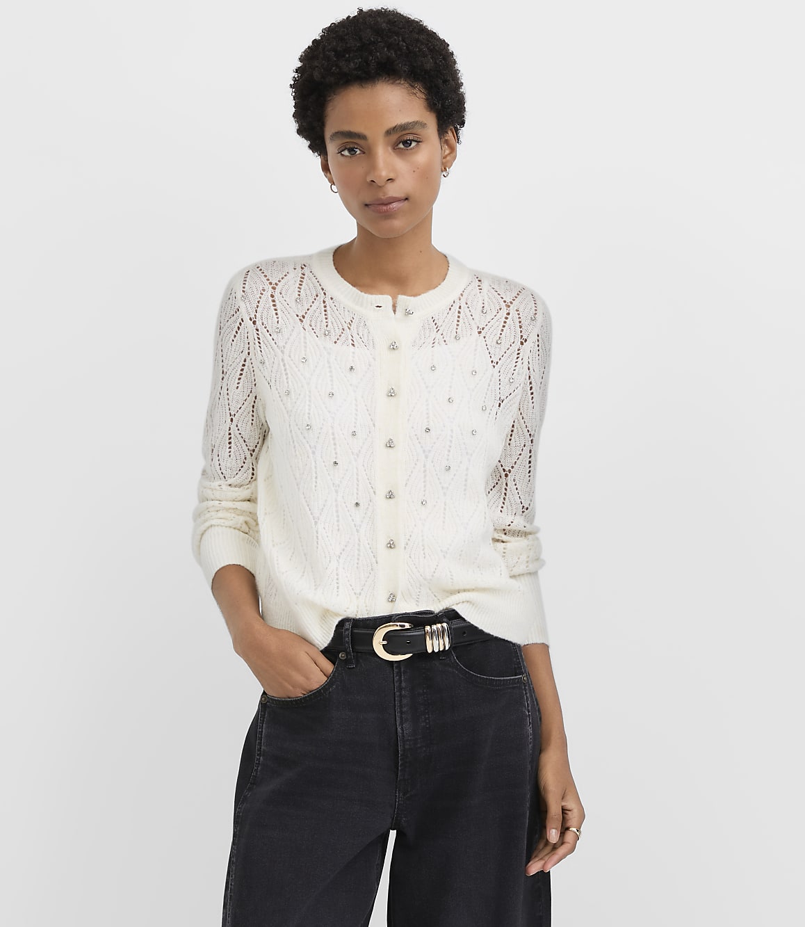 A woman wearing a white pointelle knit cardigan and black jeans stands against a plain light gray background.