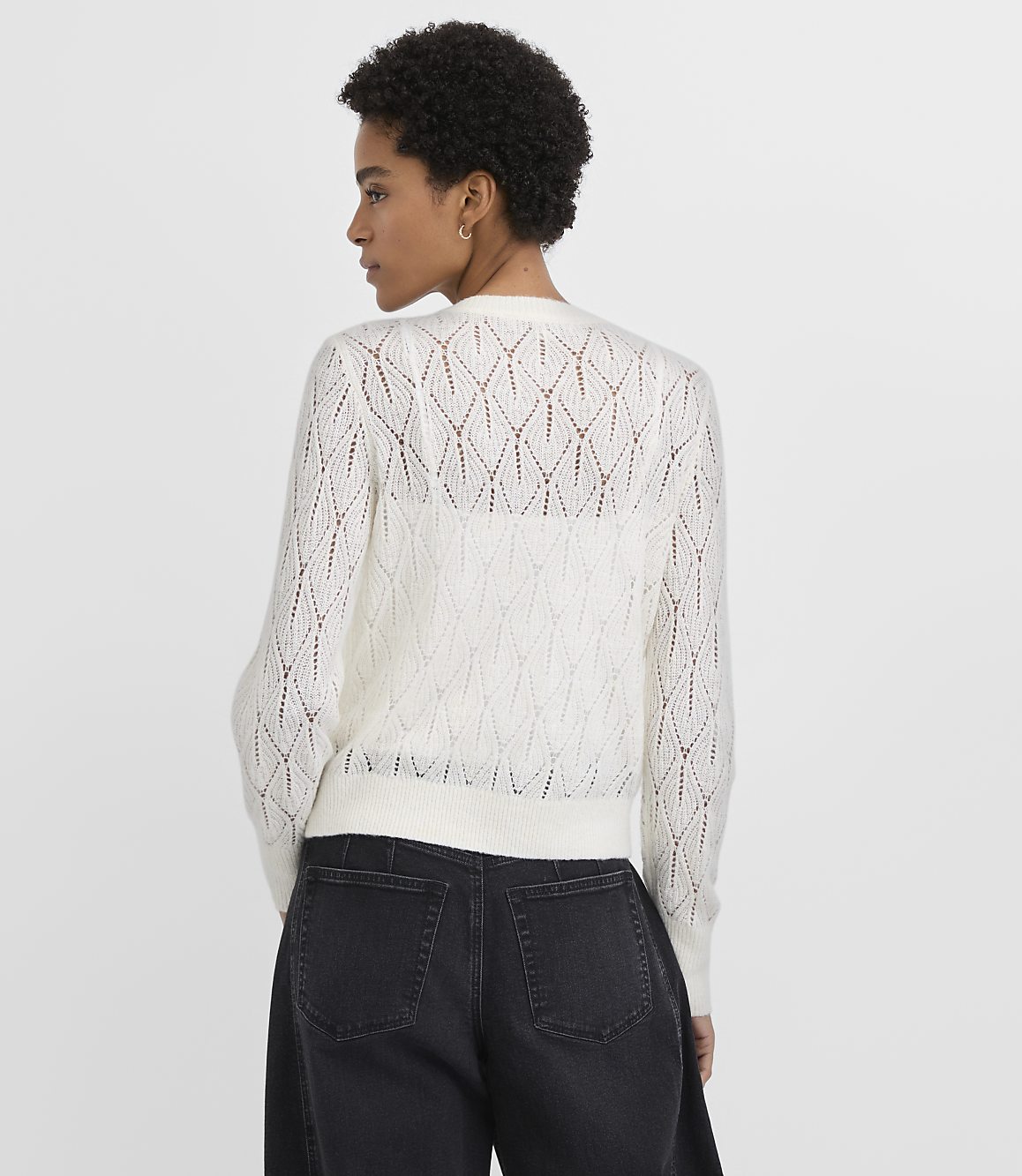 A woman wearing a white open-knit sweater and black jeans stands against a plain light gray background.