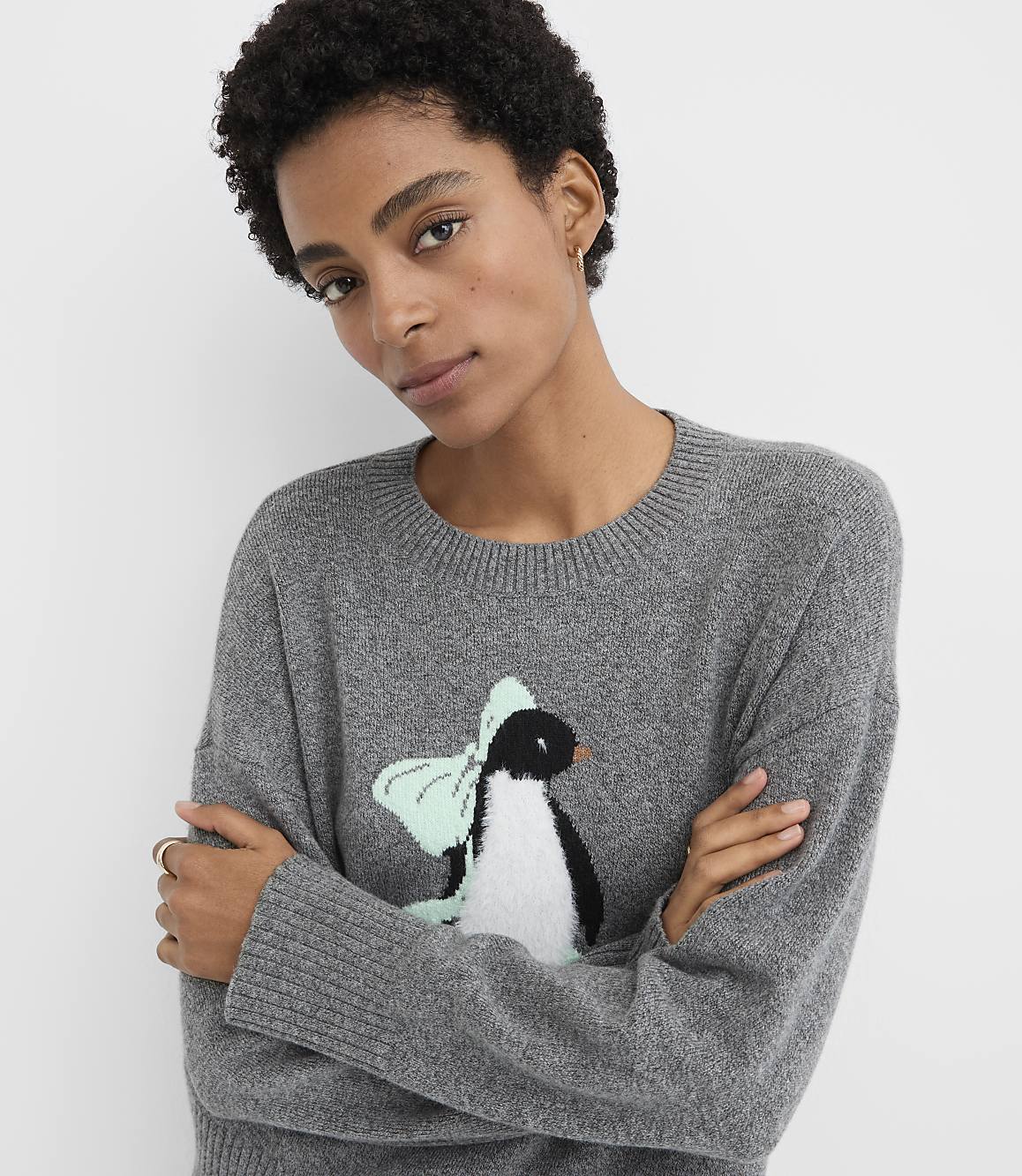 A woman with short curly hair wearing a grey sweater with a penguin graphic stands against a plain white background.