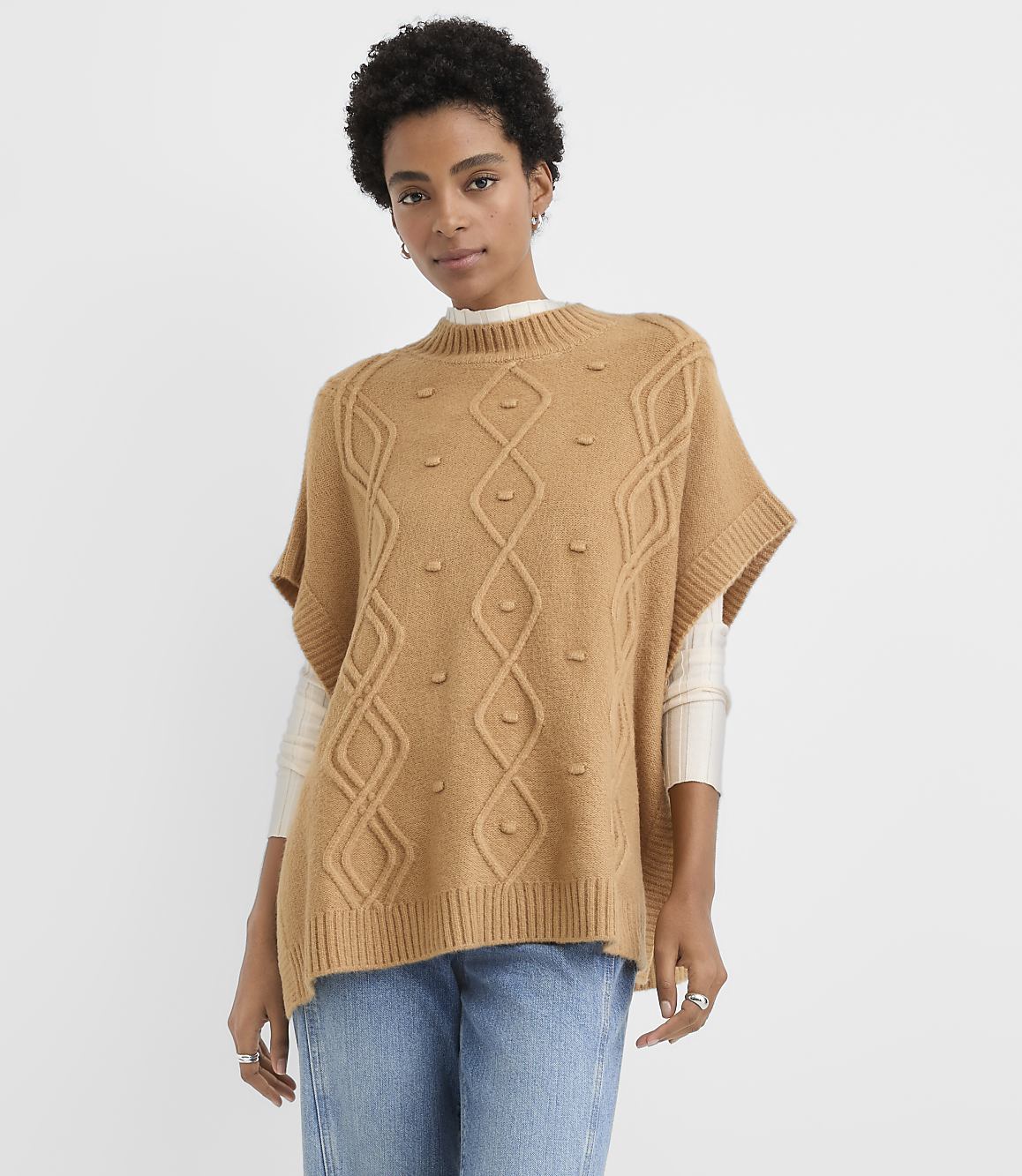 A woman wearing a tan cable-knit poncho and blue jeans stands against a plain white background.