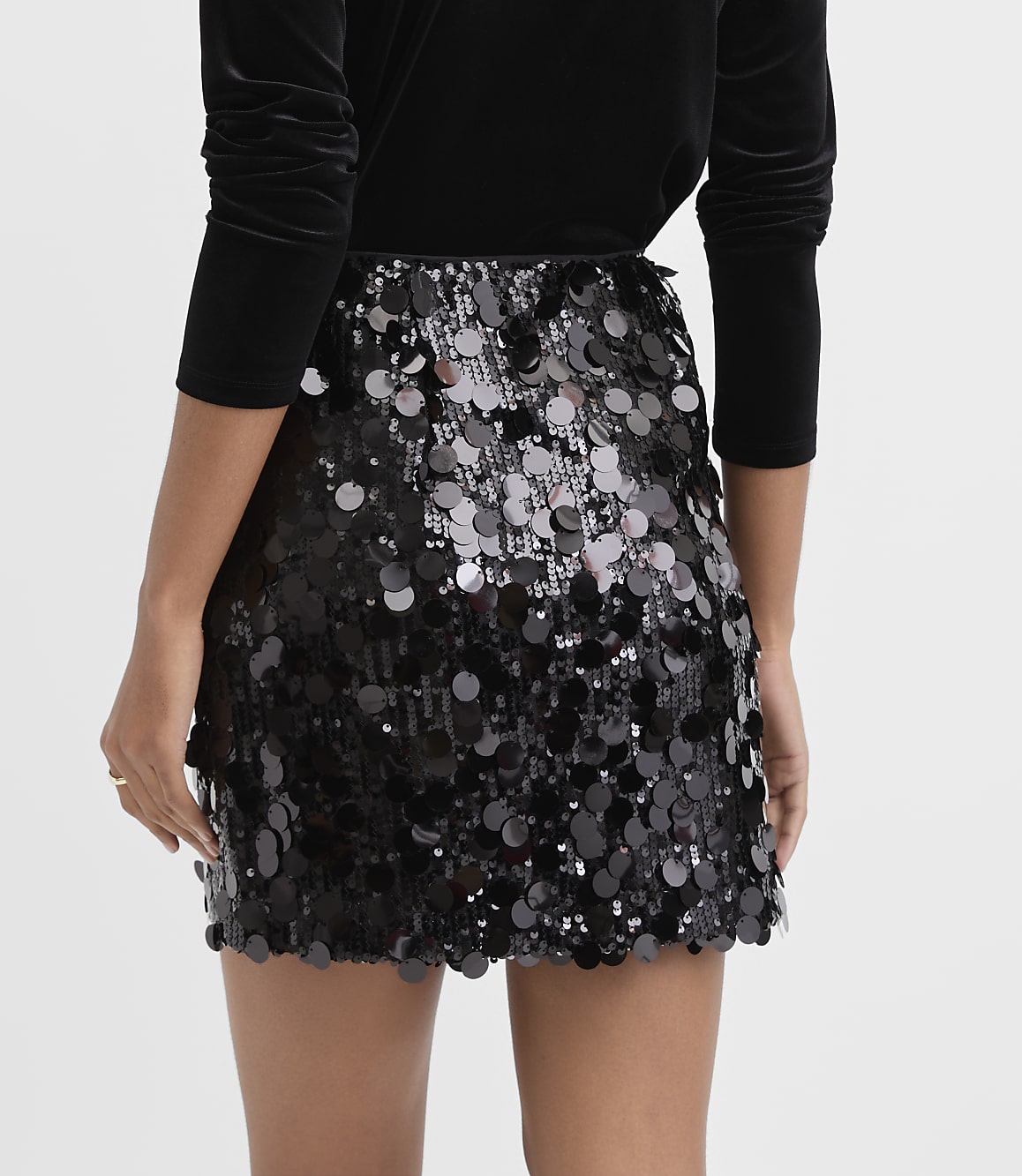 A person wears a black velvet top and a shimmering black sequined mini skirt against a plain white background.