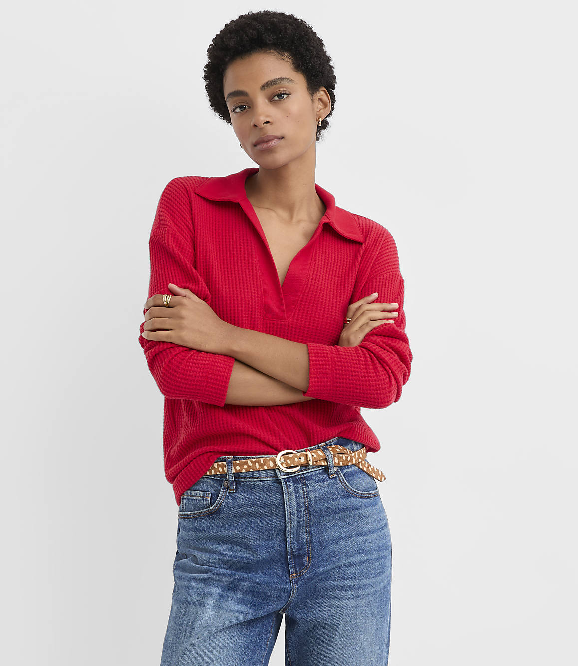 A woman wearing a red ribbed polo shirt and blue jeans stands with her arms crossed against a plain white background.