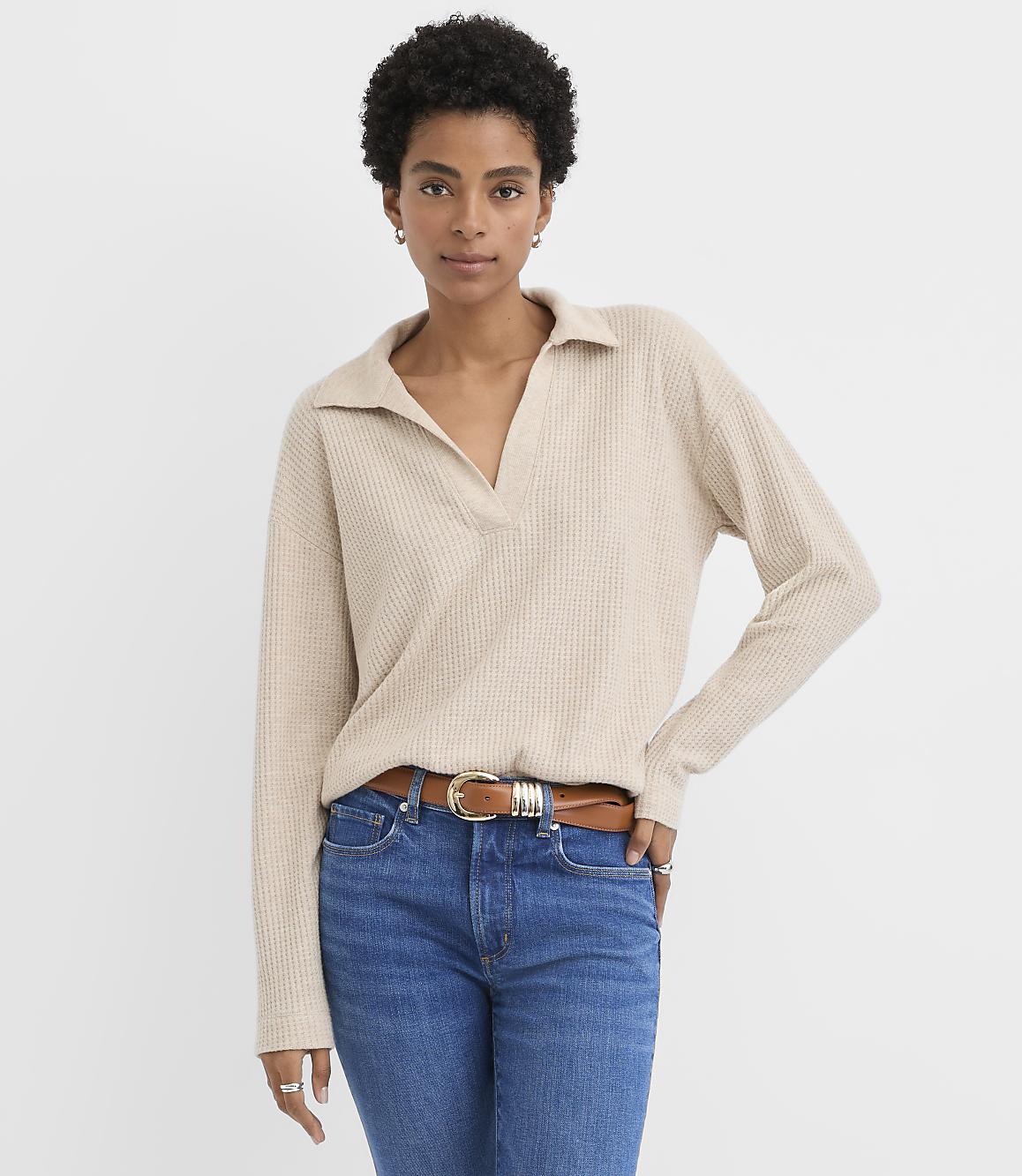 A woman wearing a beige collared sweater and blue jeans stands against a plain white background.