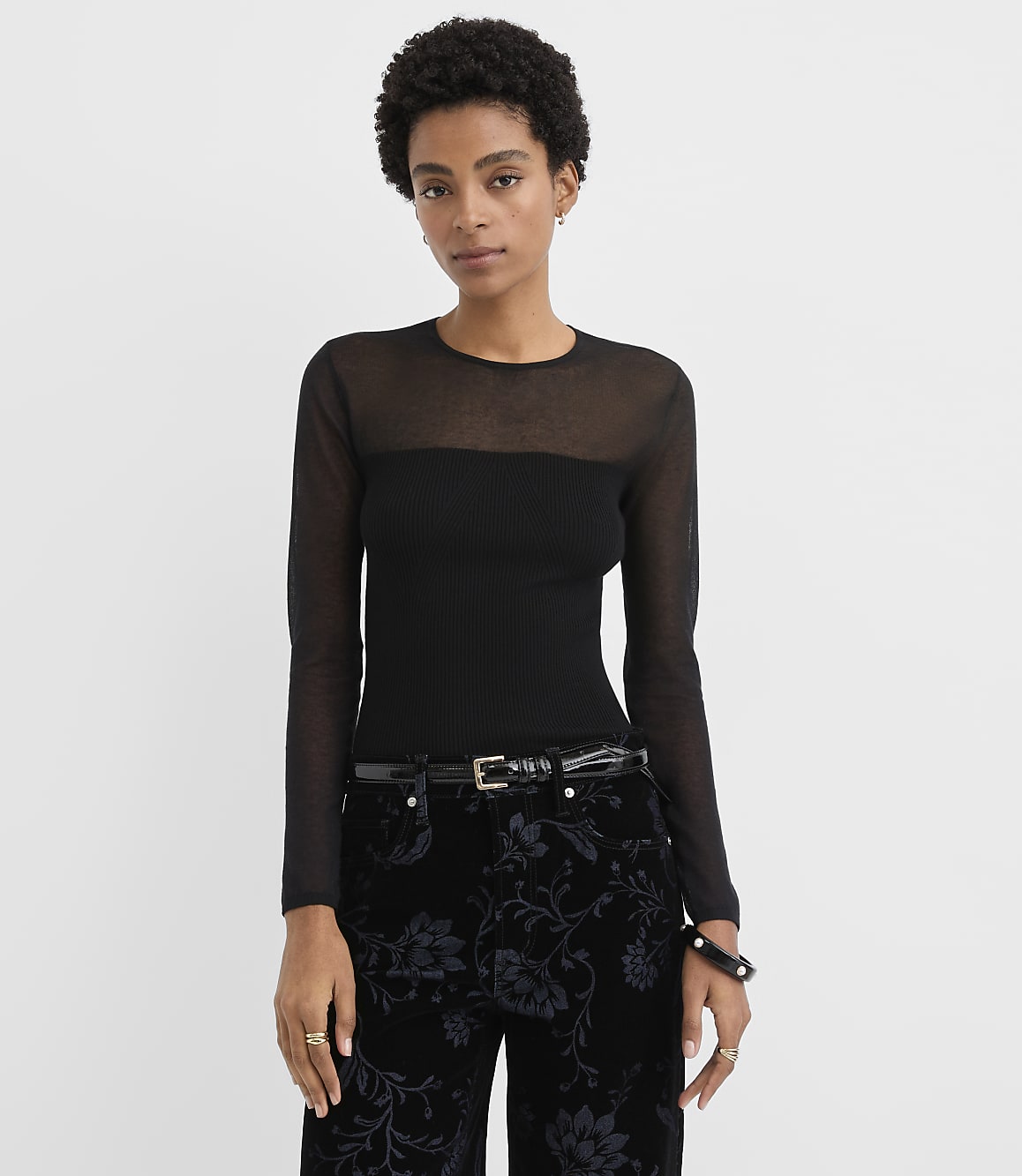 A woman wearing a sheer-sleeved black top and floral patterned black pants stands against a plain white background.
