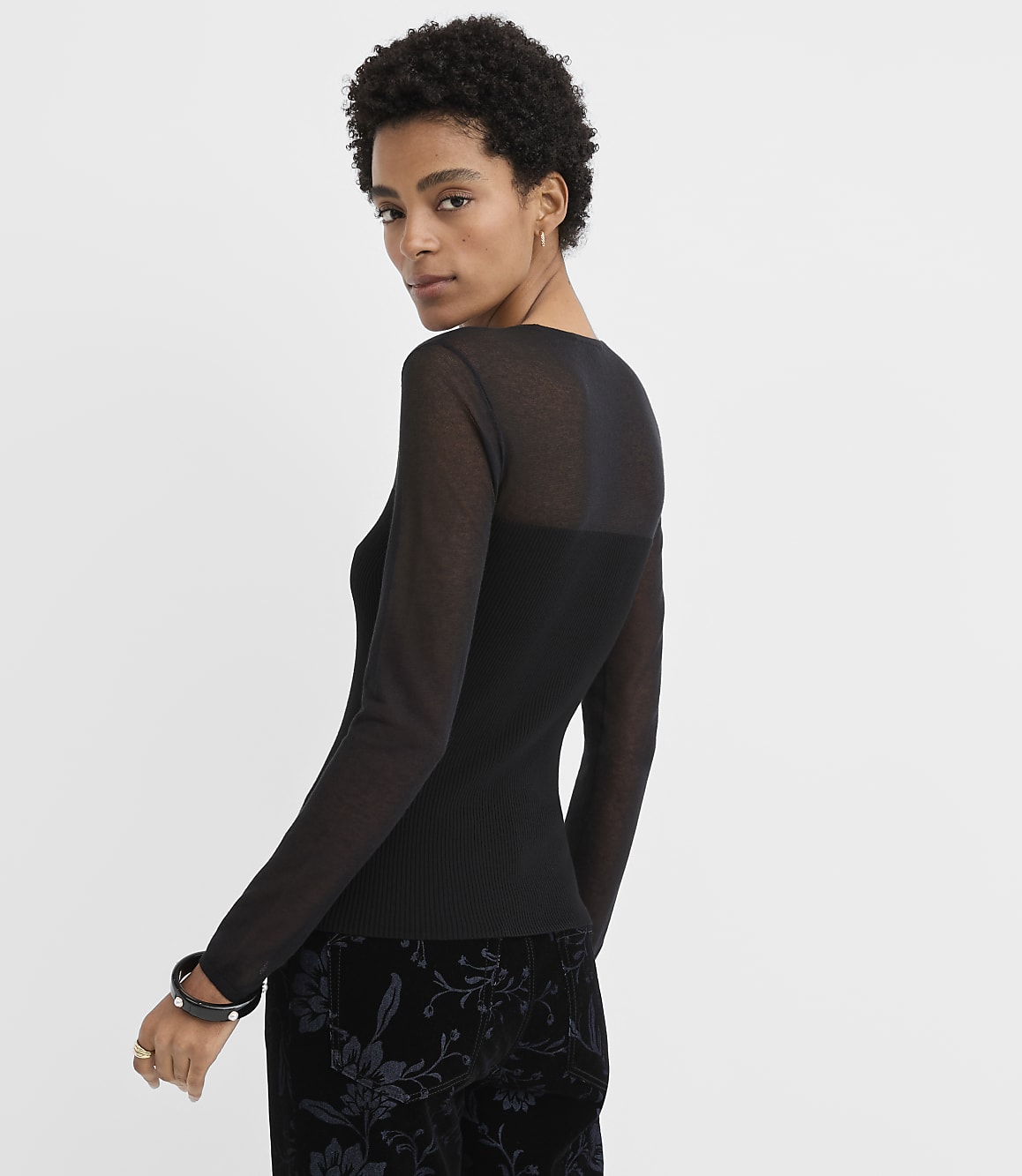A woman wearing a sheer black long-sleeved top and floral velvet trousers poses against a plain white background.
