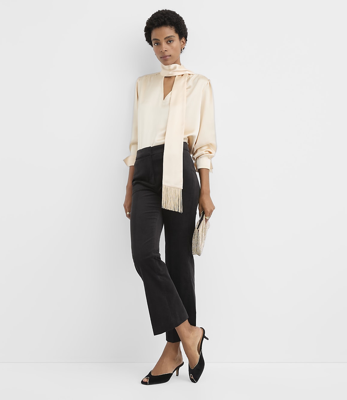 A woman wearing a champagne-colored satin blouse, a matching scarf, and black trousers stands against a plain white background.
