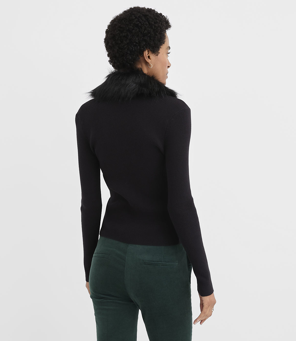 A woman wearing a black ribbed top with a faux-fur collar and dark green trousers stands against a plain white background.