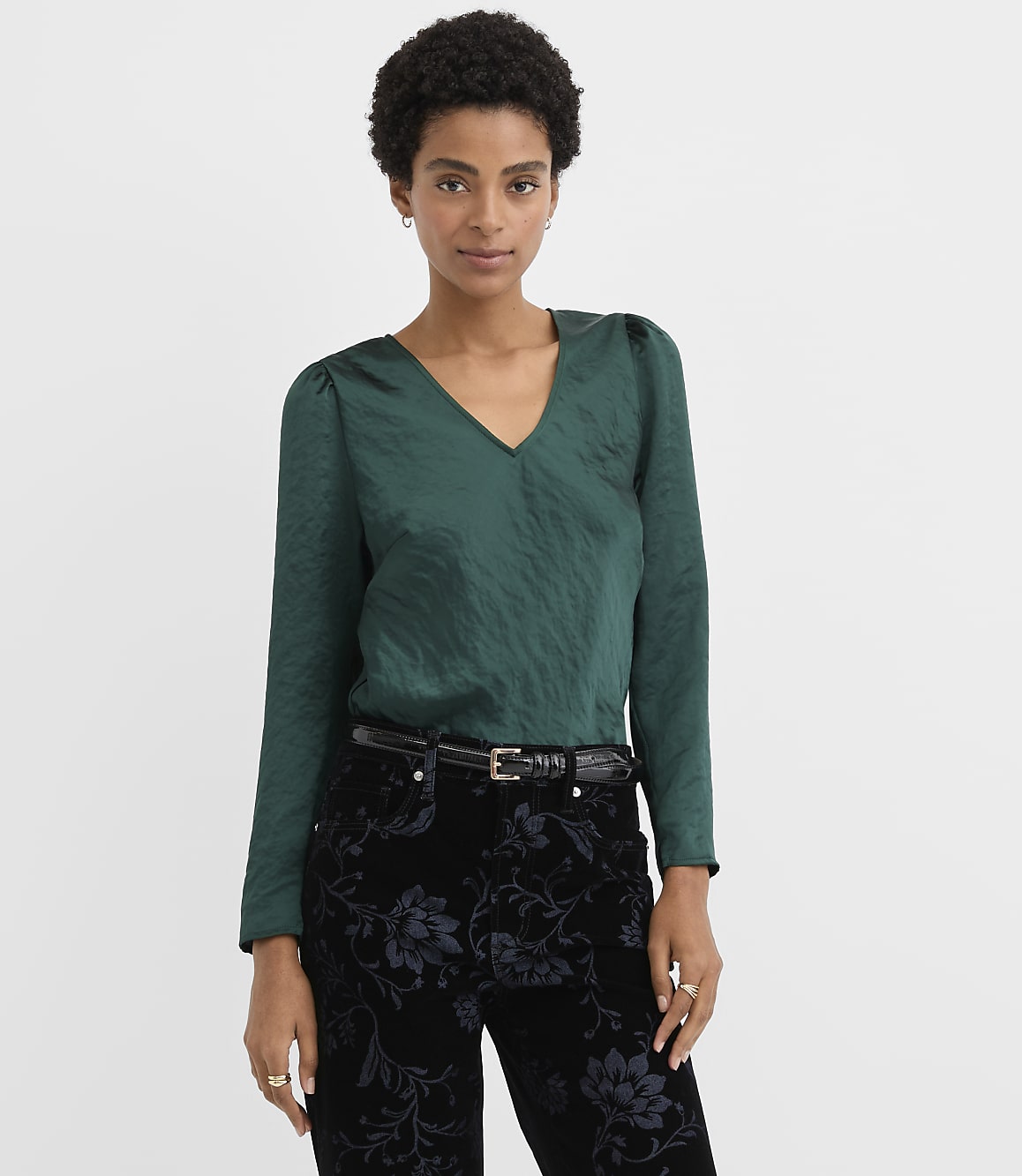 A woman wearing a dark green V-neck blouse and floral black trousers stands against a plain white background.