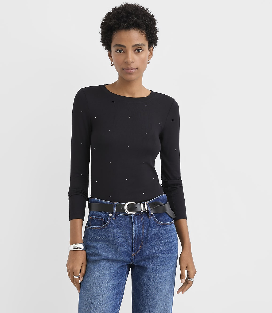 A woman wearing a black studded top and blue jeans stands against a plain white background.