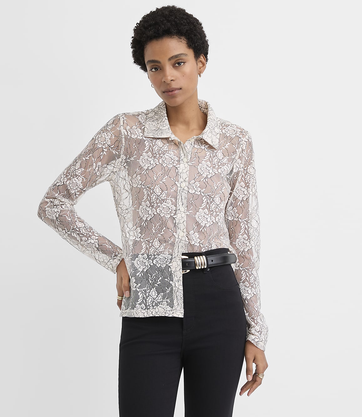 A woman wearing a sheer white lace button-down shirt and black trousers stands against a plain white background.