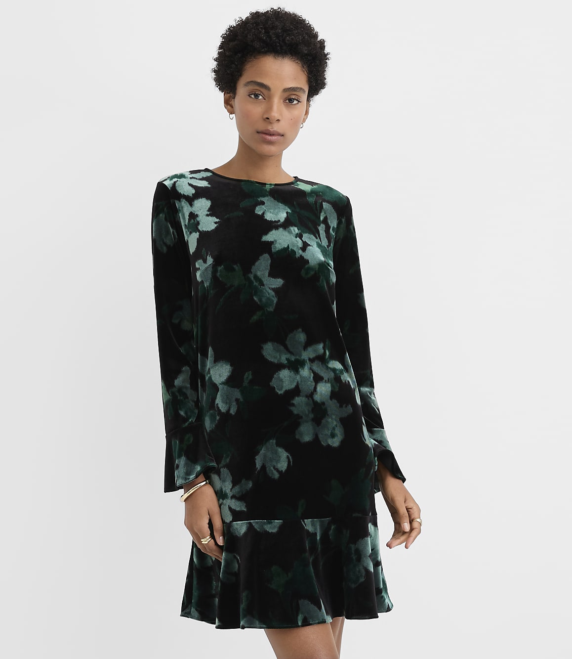A woman wearing a black and green floral velvet dress stands against a plain white background.
