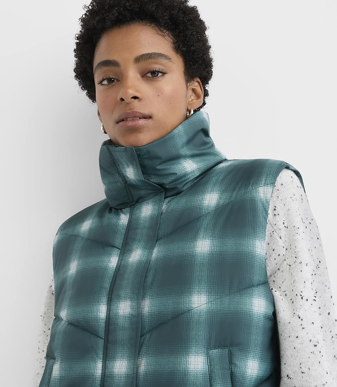 A woman wearing a green plaid puffer vest over a white speckled sweater stands against a plain white background.