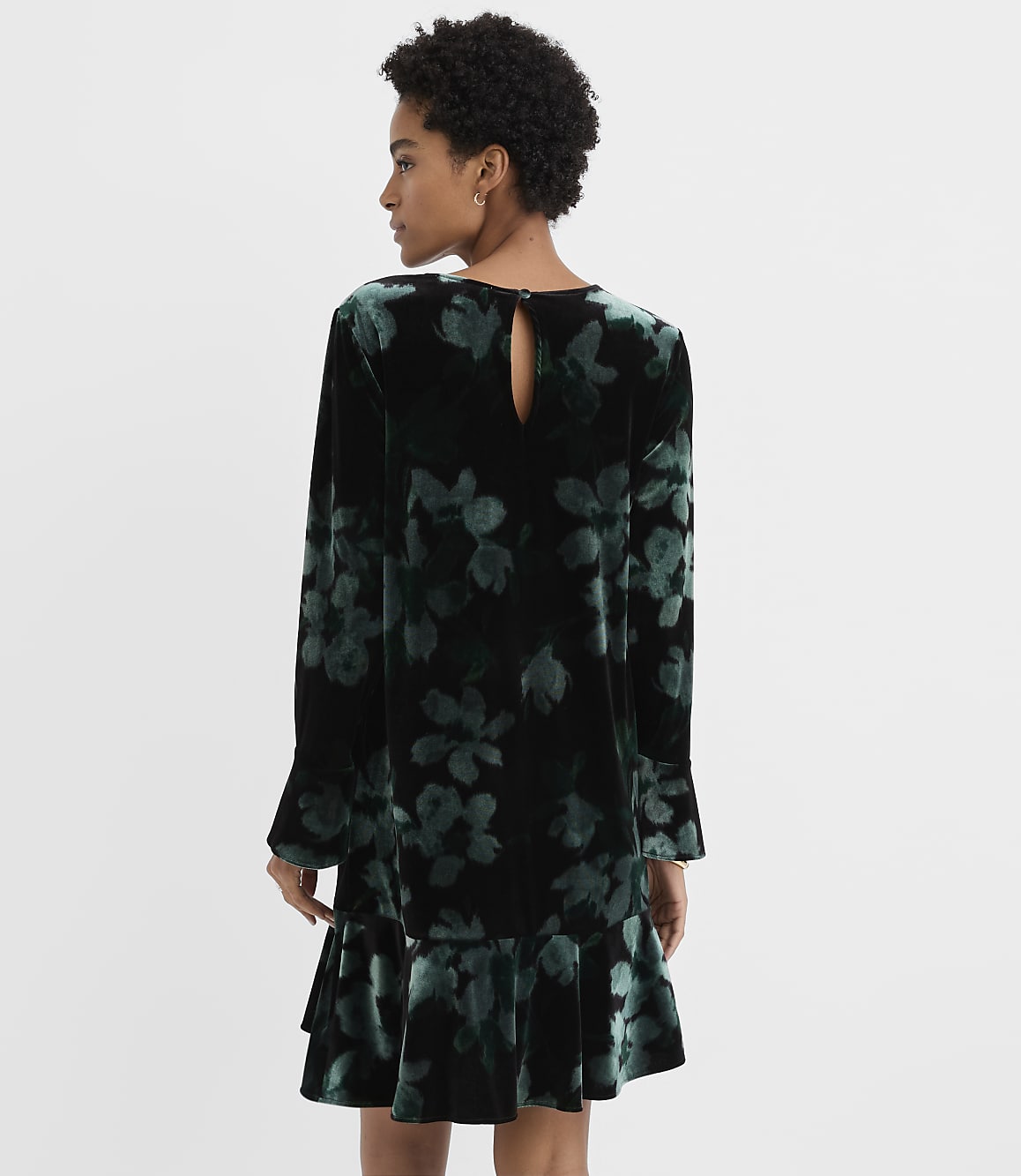 A woman stands with her back to the camera wearing a dark green floral velvet dress against a plain white background.