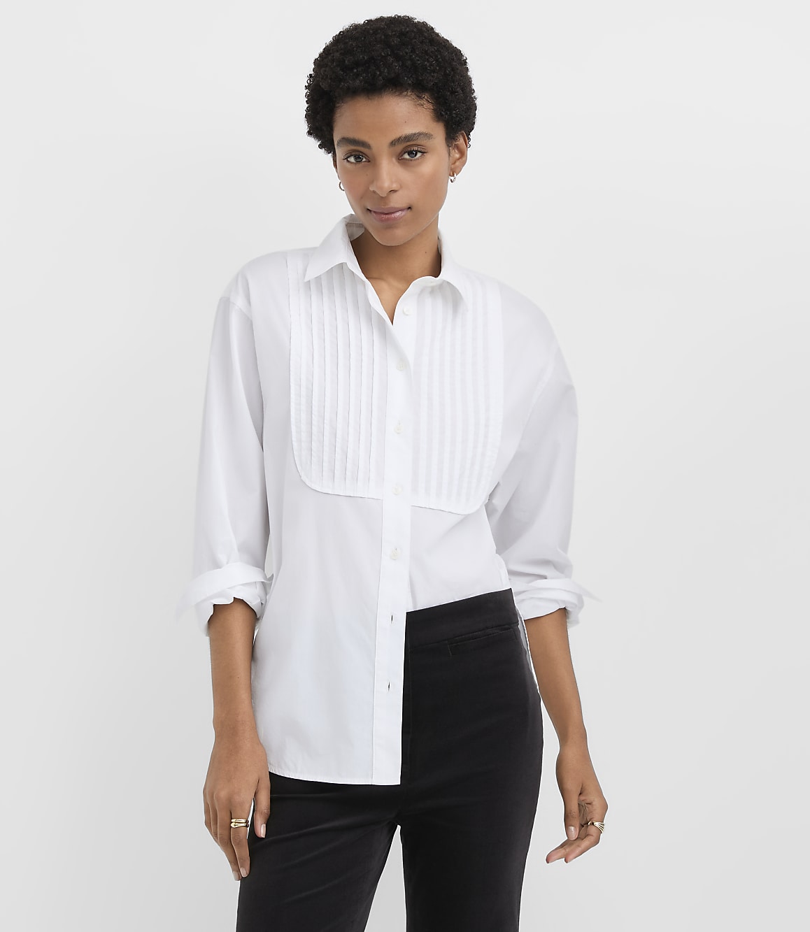 A woman wearing a white pleated button-down shirt and black trousers stands against a plain white background.