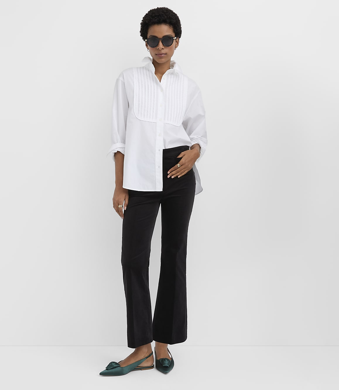 A woman wearing a white pleated shirt, black trousers, and green flats stands against a plain white background.