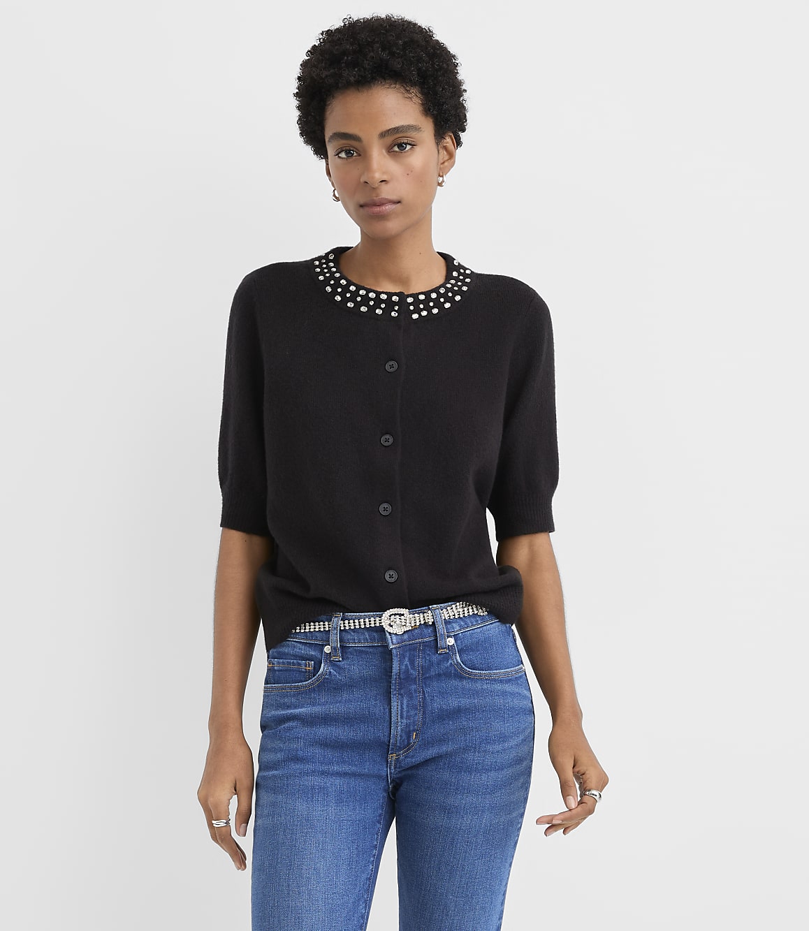 A woman wearing a black embellished cardigan and blue jeans stands against a plain light gray background.