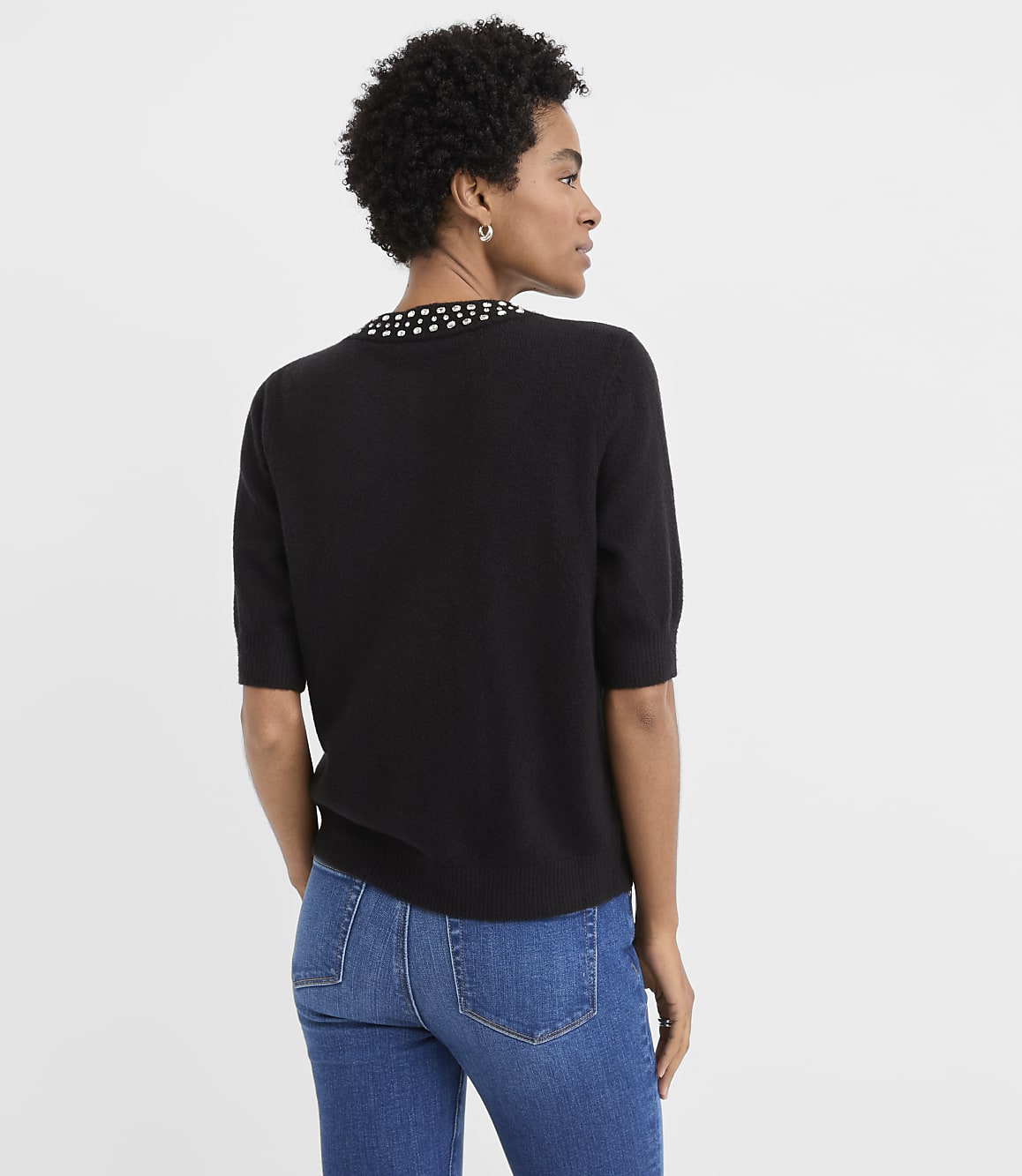 A woman wearing a black short-sleeved sweater with a beaded collar and blue jeans stands against a plain white background.