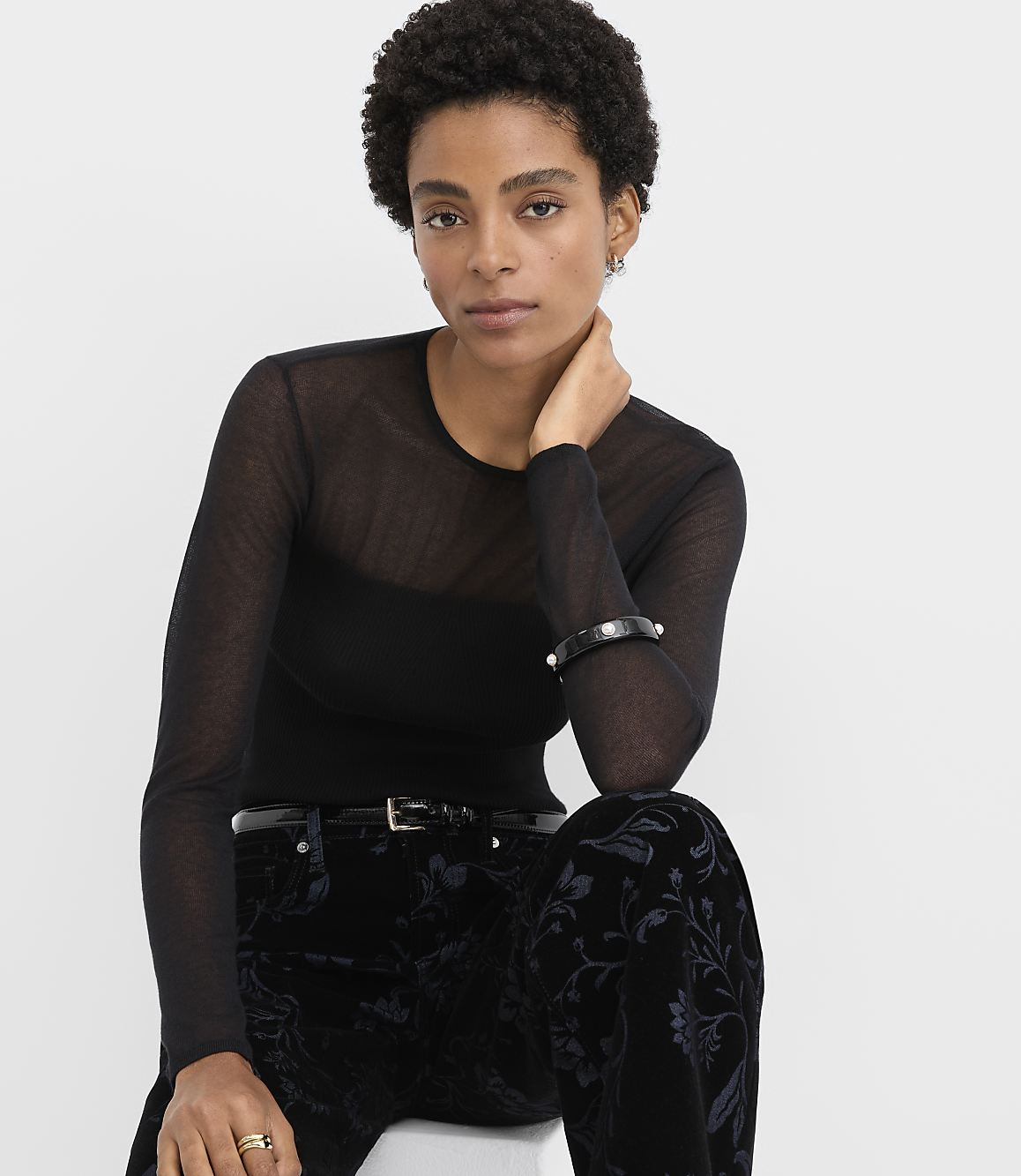 A woman wearing a sheer black long-sleeved top and floral velvet trousers poses against a plain white background.