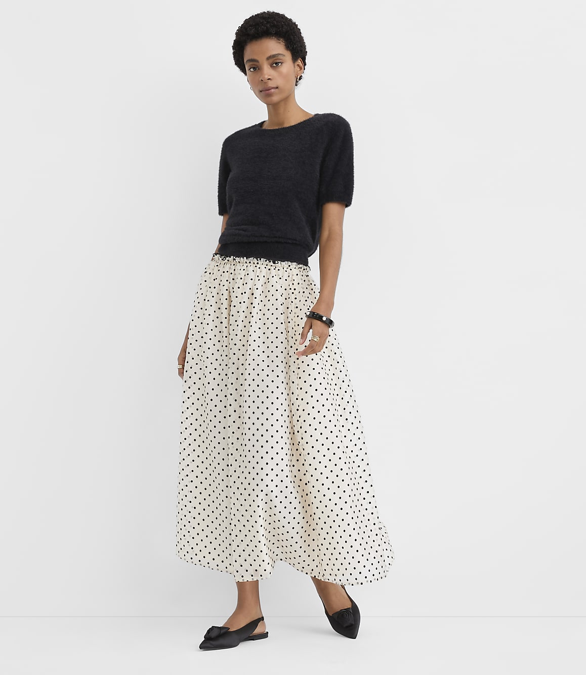 A woman wearing a black short-sleeved top and a long white polka-dot skirt stands against a plain white background.
