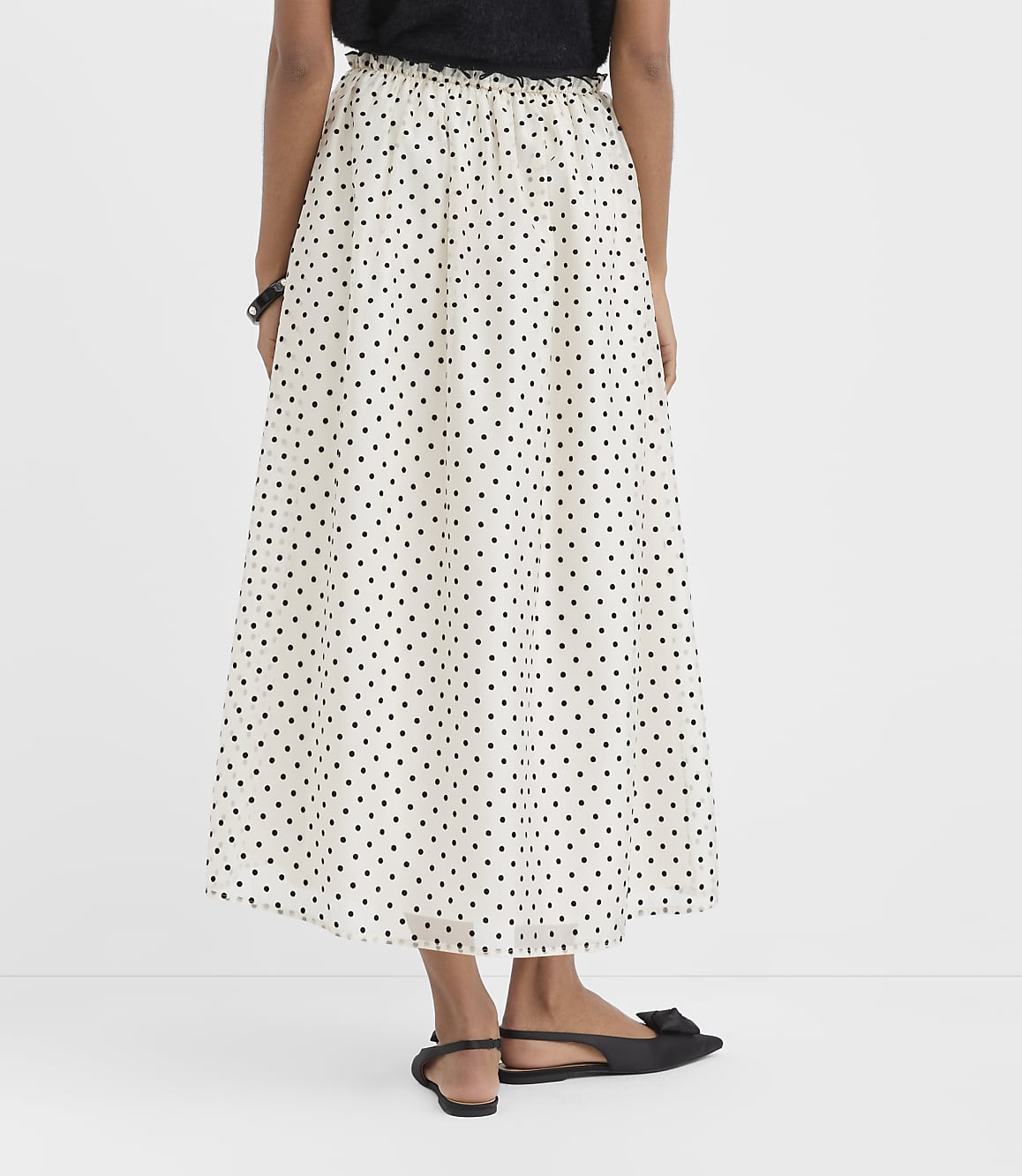A person wearing a black top and a white polka-dot maxi skirt with black flats stands against a plain white background.