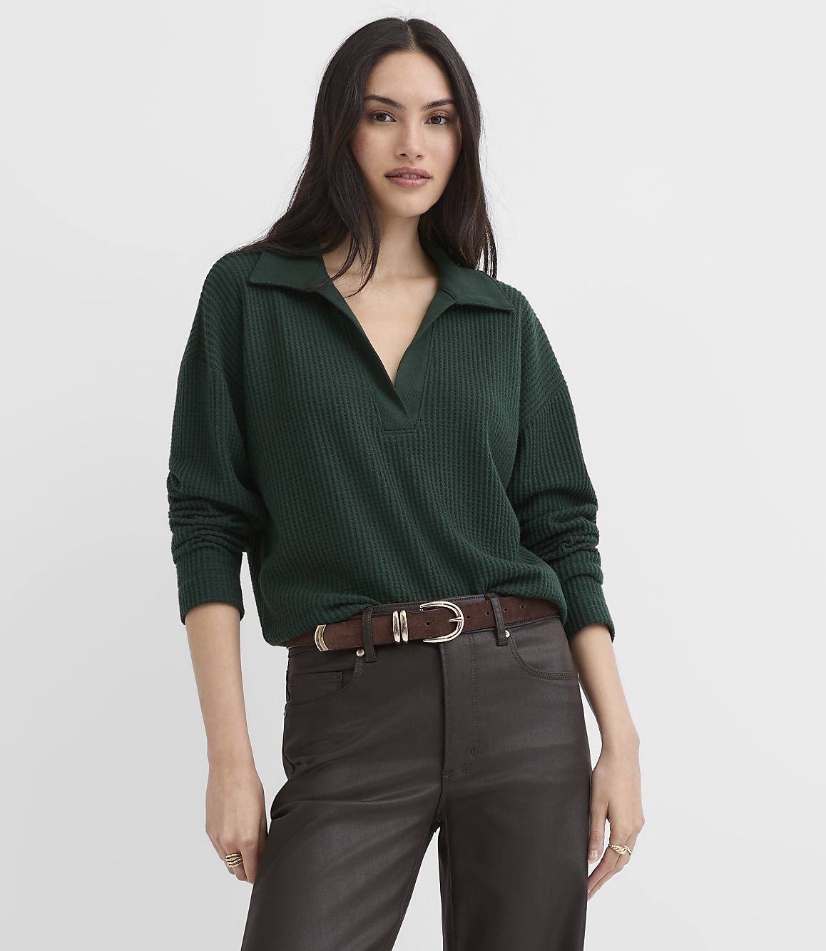 A woman wearing a dark green ribbed collared top and brown leather pants stands against a plain white background.