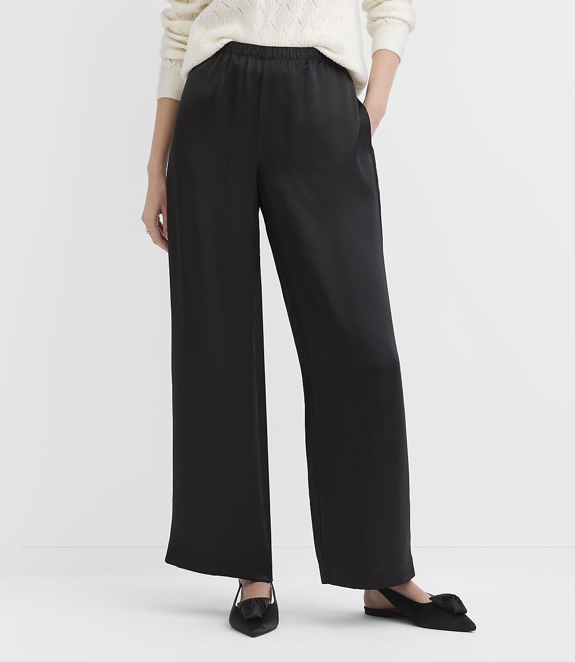 A person wears wide-leg black trousers, a white sweater, and black pointed flats against a plain white background.