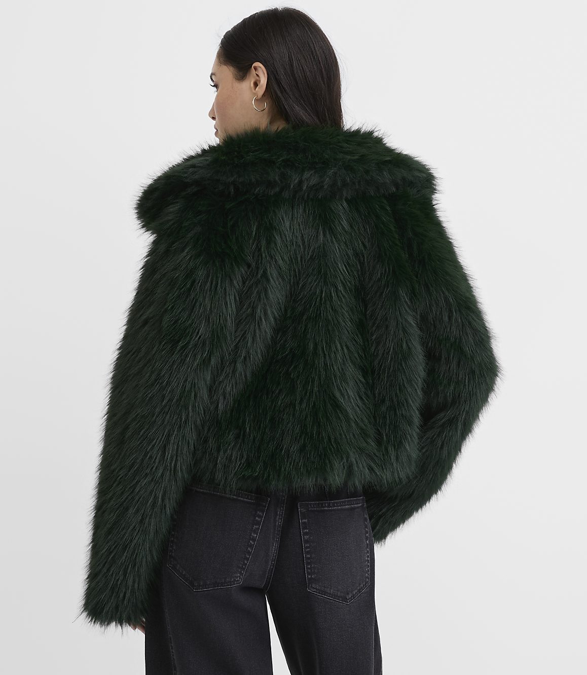 A woman wearing a dark green faux fur coat and black jeans stands against a plain white background.