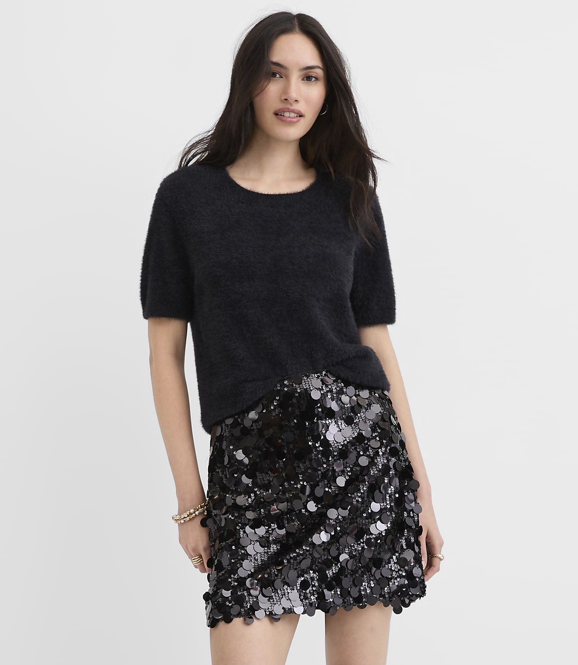 A woman wearing a fuzzy black short-sleeved sweater and a sequined black mini skirt stands against a plain white background.