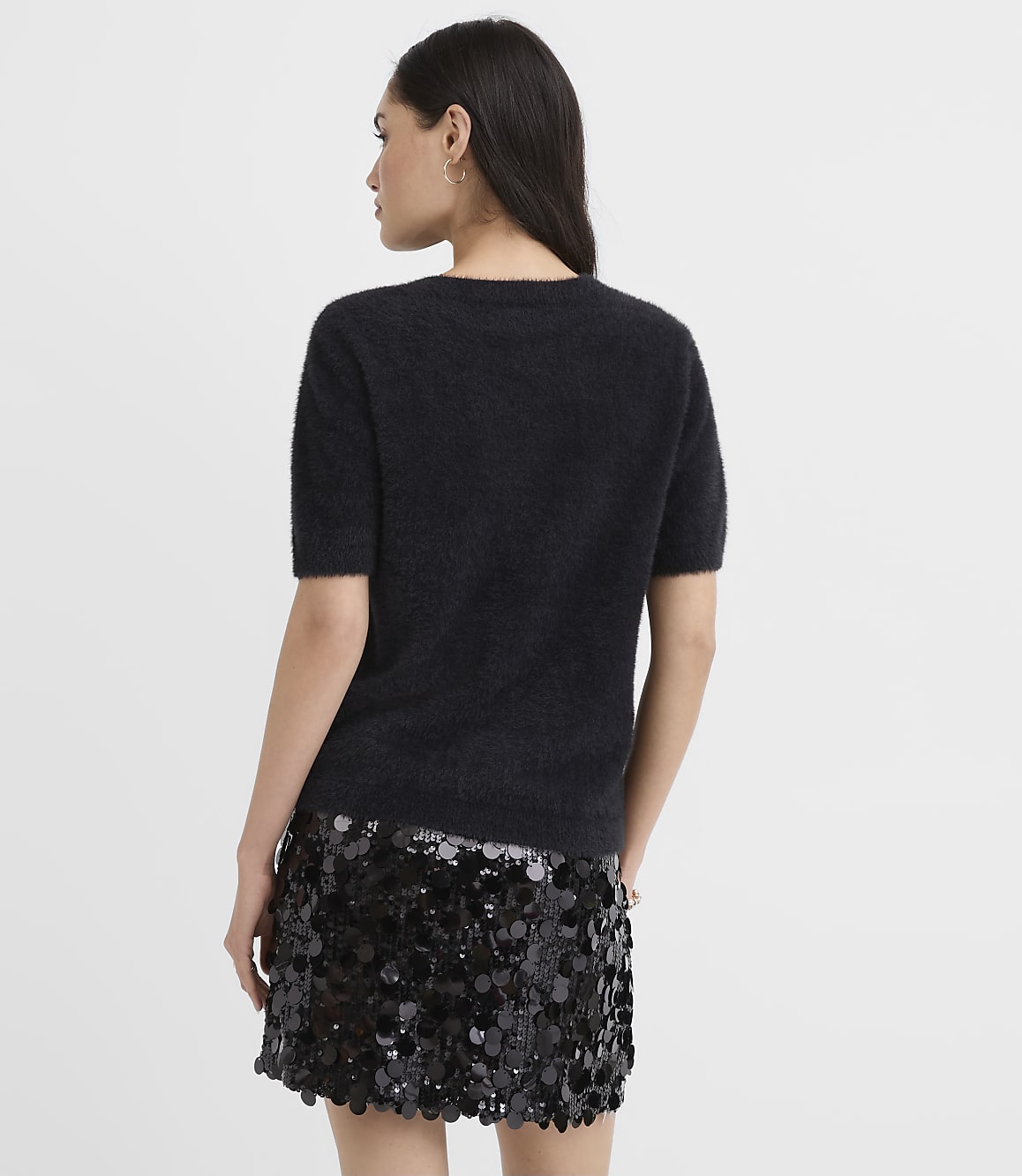 A woman wearing a fuzzy black short-sleeved top and a sequined black skirt stands with her back to the camera against a plain light gray background.