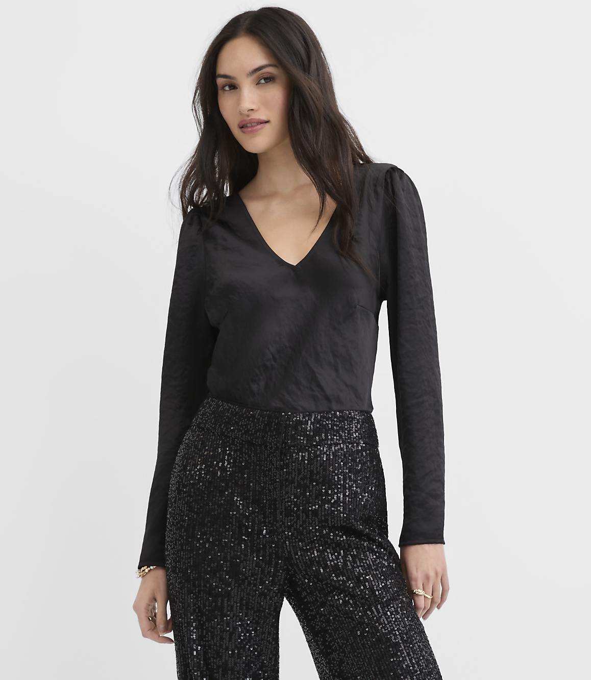 A woman wearing a black V-neck top and sequined black pants stands against a plain white background.