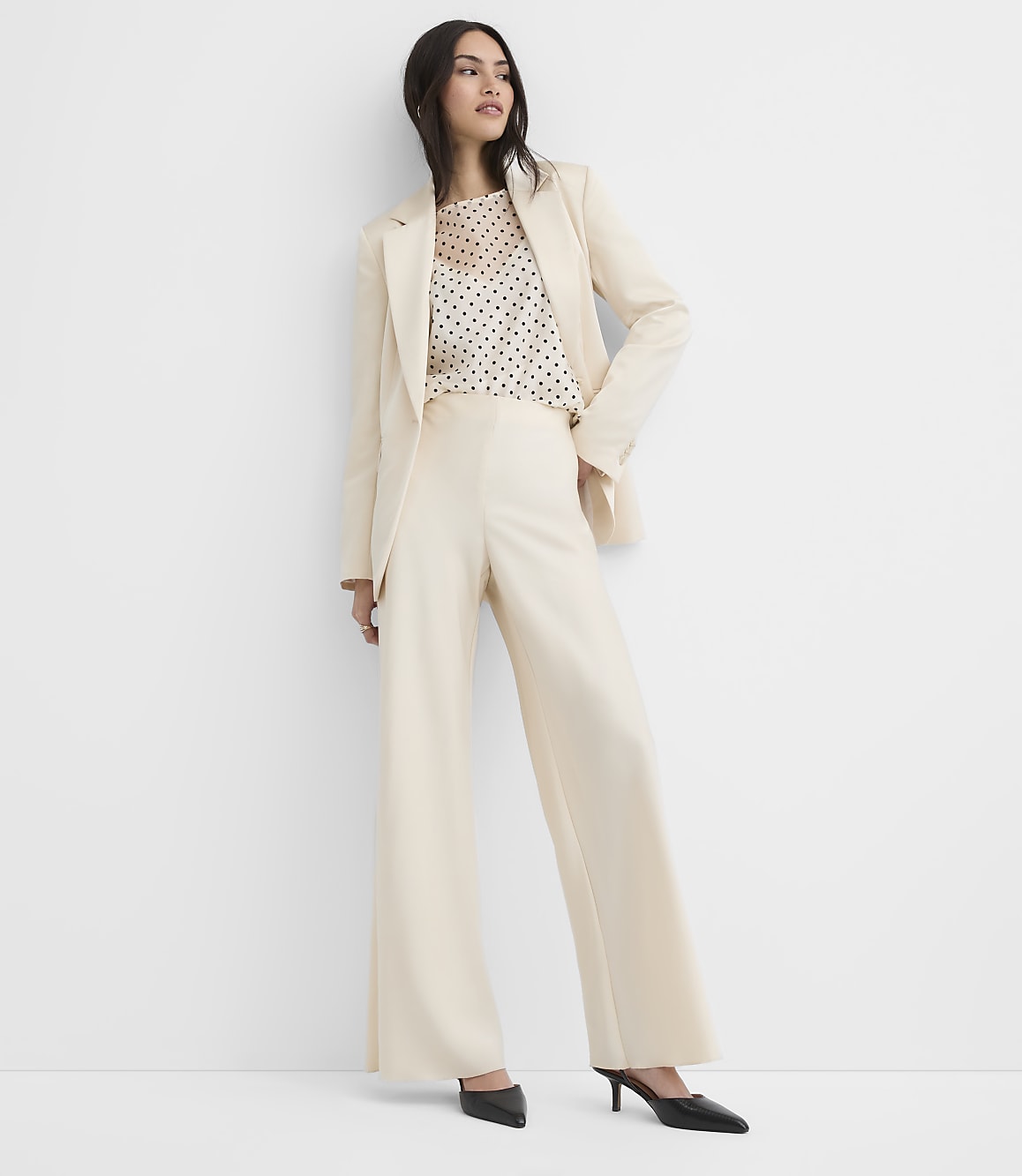 A woman wearing a cream-colored suit and a polka-dot top stands against a plain white background.
