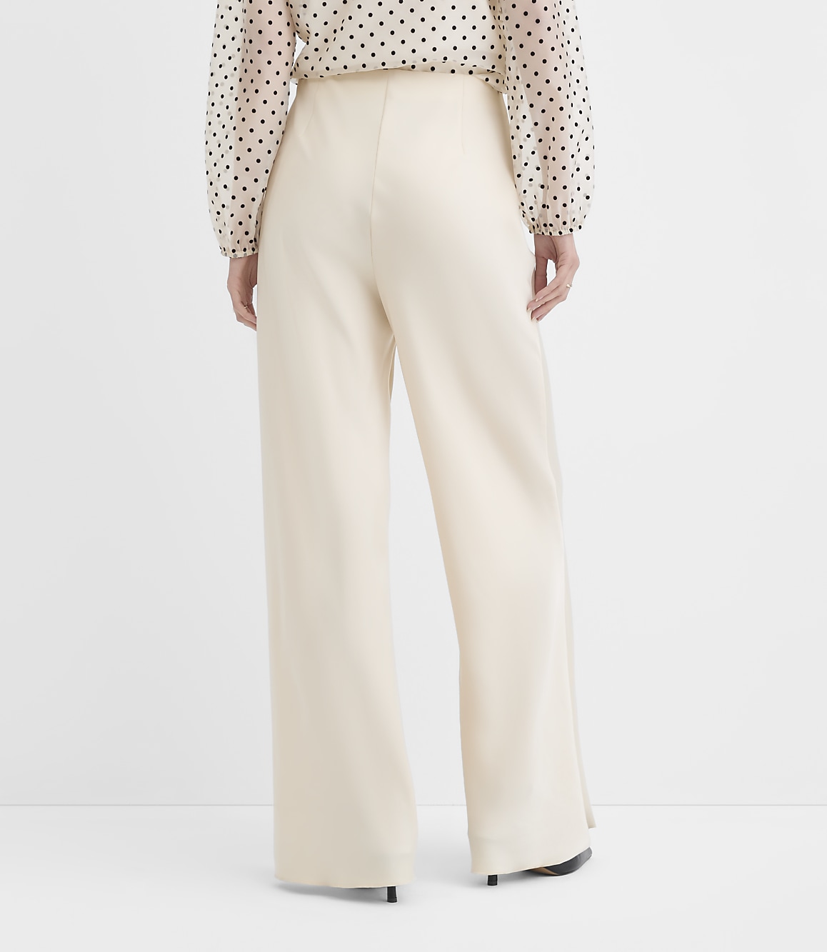 A person wearing a polka-dot blouse and wide-leg cream trousers stands against a plain white background.