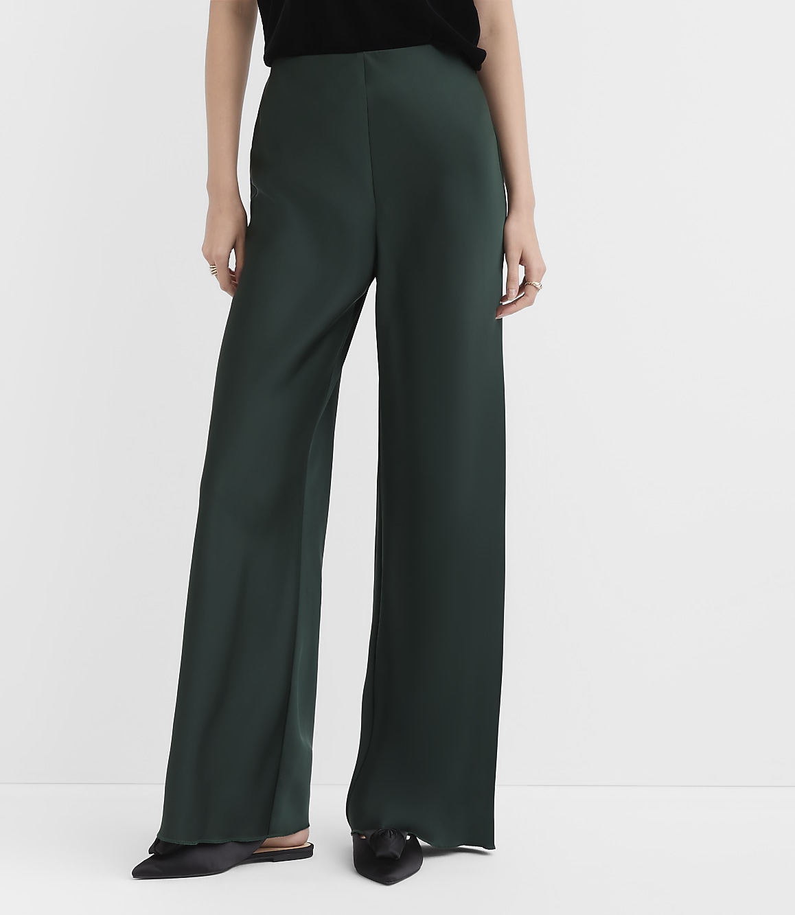 A person wearing a black top, dark green wide-leg trousers, and black pointed-toe flats stands against a plain white background.