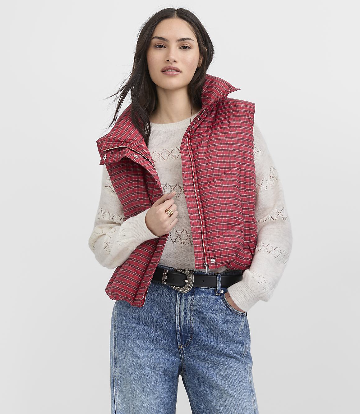 A woman wearing a red plaid puffer vest, a cream sweater, and blue jeans stands against a plain white background.