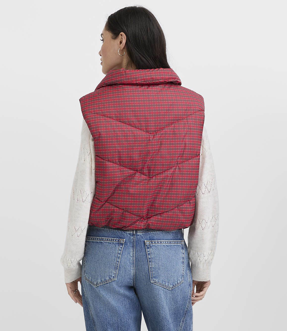 A woman wearing a red plaid puffer vest, a white sweater, and blue jeans stands against a plain light gray background.