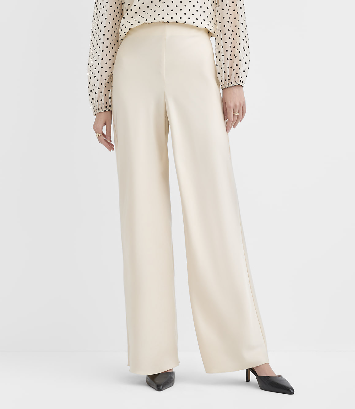 A person wears a polka-dot blouse and wide-leg cream trousers with black pointed heels against a plain white background.